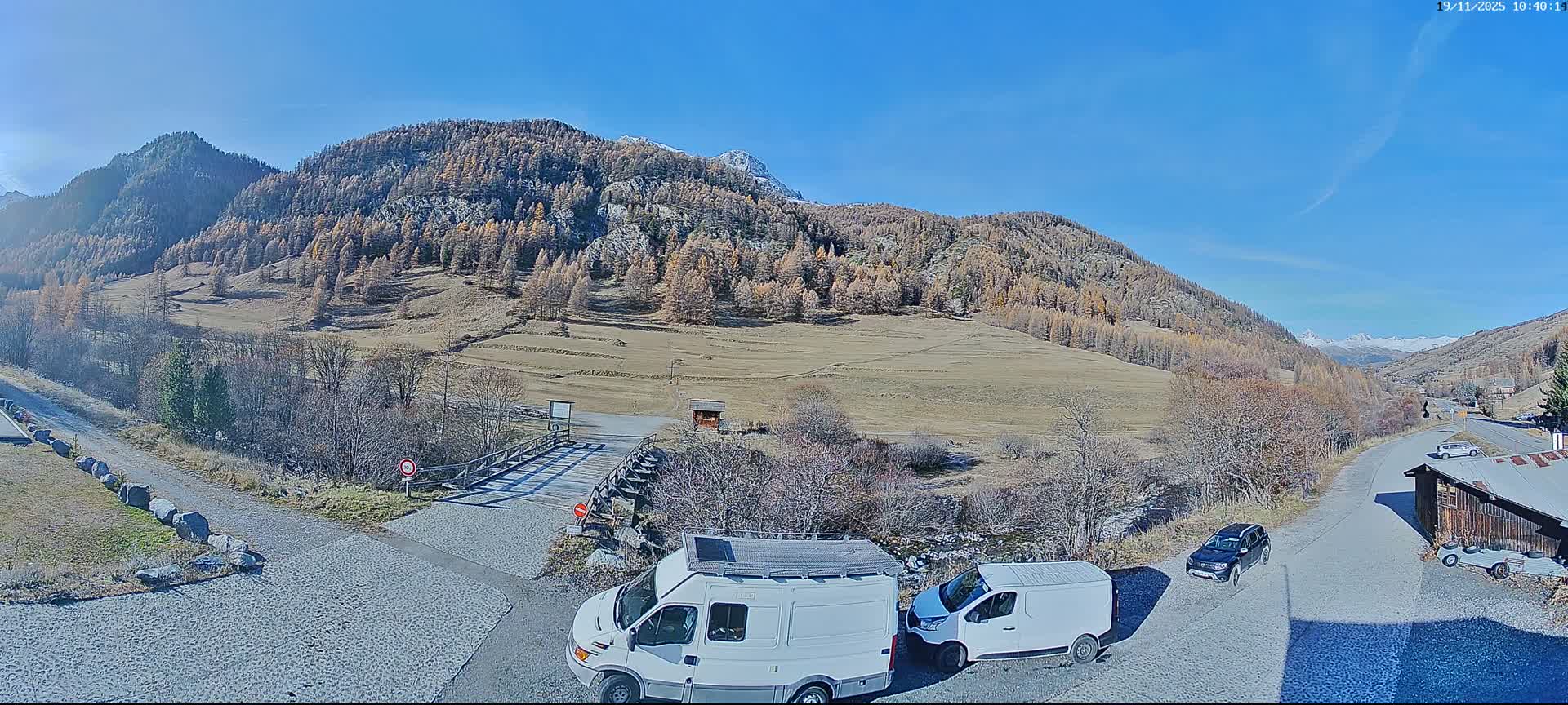 Under a clear blue sky, a sunny mountain valley features hillsides dotted with autumn-colored trees and dry grasslands, with a winding road, a wooden bridge, and several parked vehicles in the foreground.