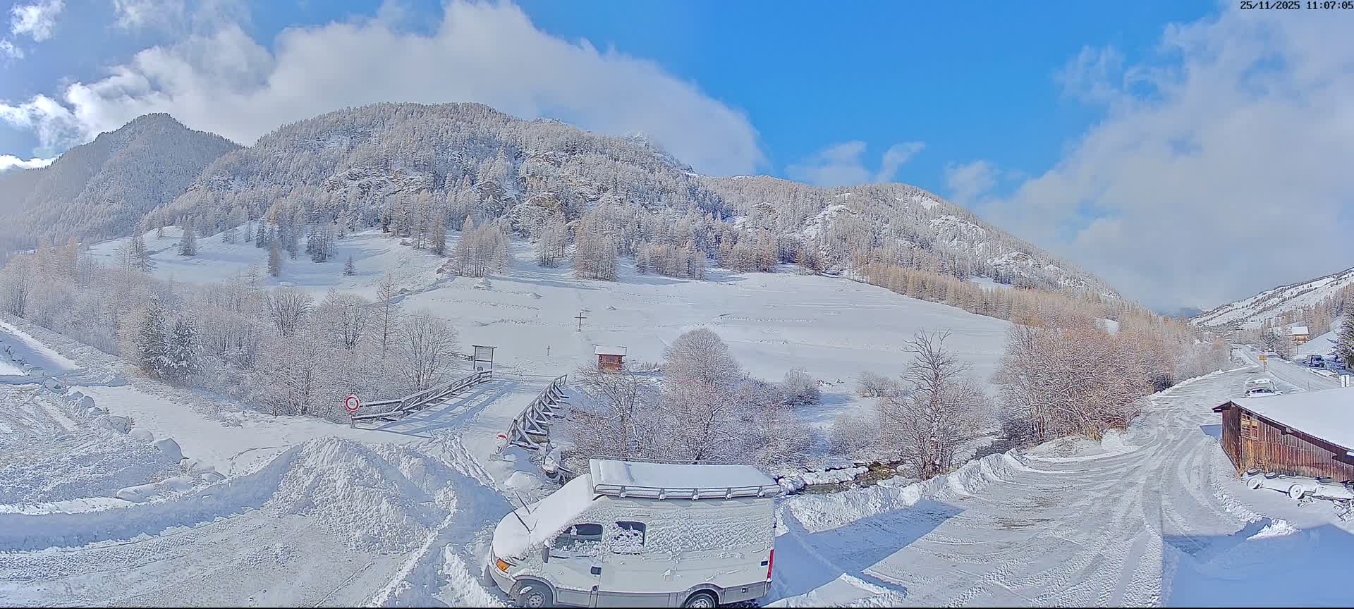 A picturesque snow-covered mountain valley features a camper van in the foreground, a winding road, a small wooden hut, and a bridge, all under a clear blue sky with scattered white clouds.
