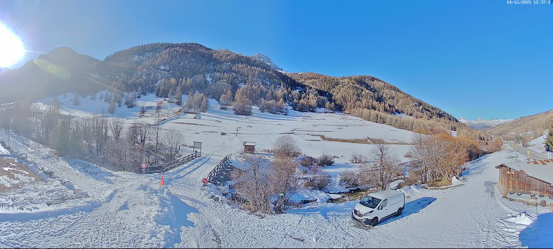A picturesque snow-covered mountain valley features a camper van in the foreground, a winding road, a small wooden hut, and a bridge, all under a clear blue sky with scattered white clouds.