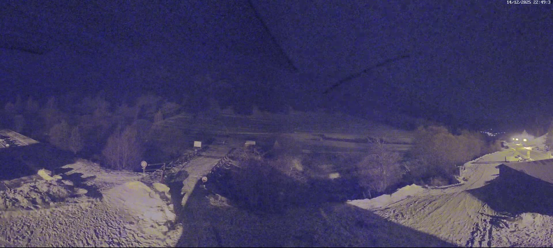 A picturesque snow-covered mountain valley features a camper van in the foreground, a winding road, a small wooden hut, and a bridge, all under a clear blue sky with scattered white clouds.