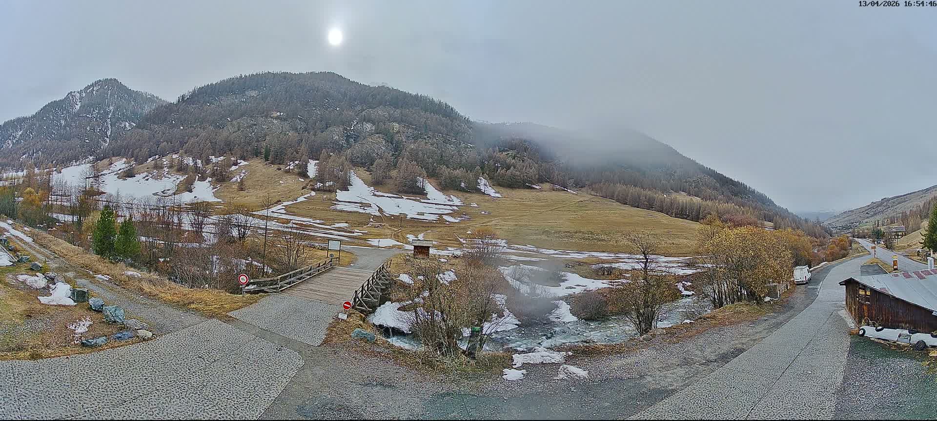 A picturesque snow-covered mountain valley features a camper van in the foreground, a winding road, a small wooden hut, and a bridge, all under a clear blue sky with scattered white clouds.
