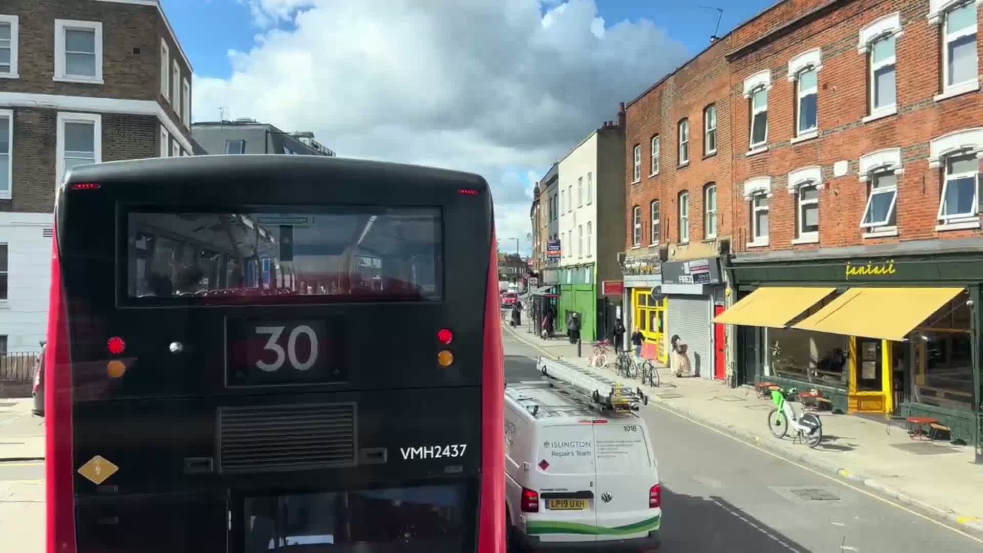 The rear of a double-decker bus numbered 30 is seen driving down a street of brick buildings on a sunny day with partly cloudy skies.