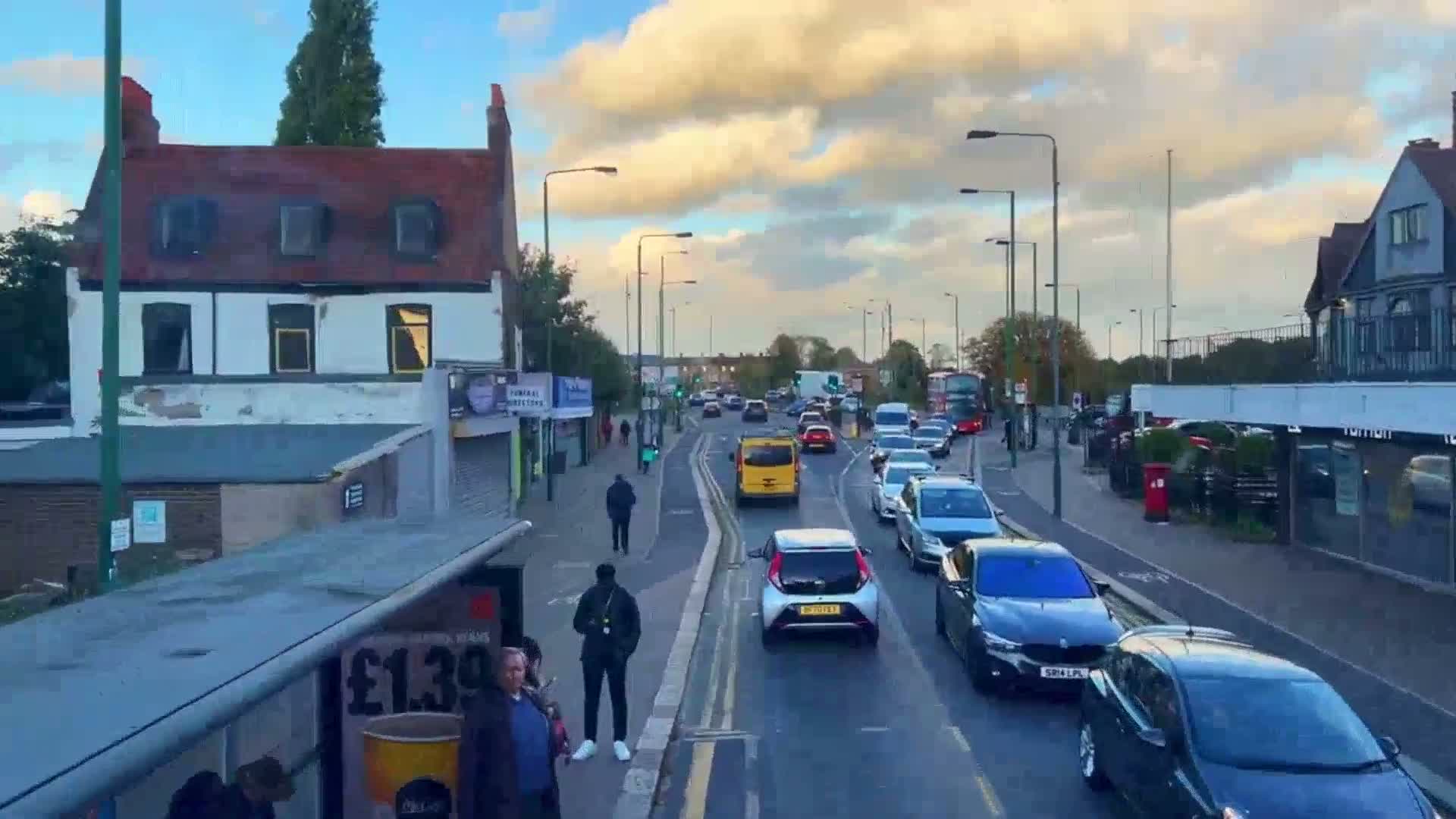 A partly cloudy evening scene shows a street with moderate traffic congestion, pedestrians on a sidewalk, and buildings lining the road.