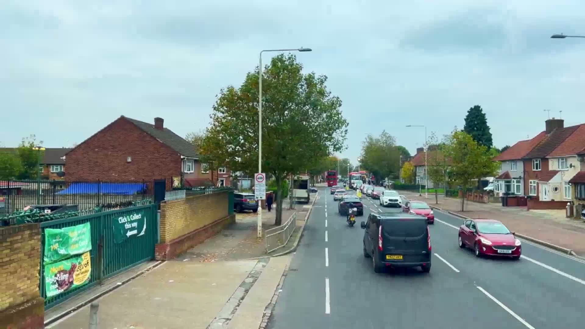 A residential street with two-story houses on either side, under an overcast sky, shows several cars and a van driving on a two-lane road, with a motorcyclist and a bus visible in the distance.