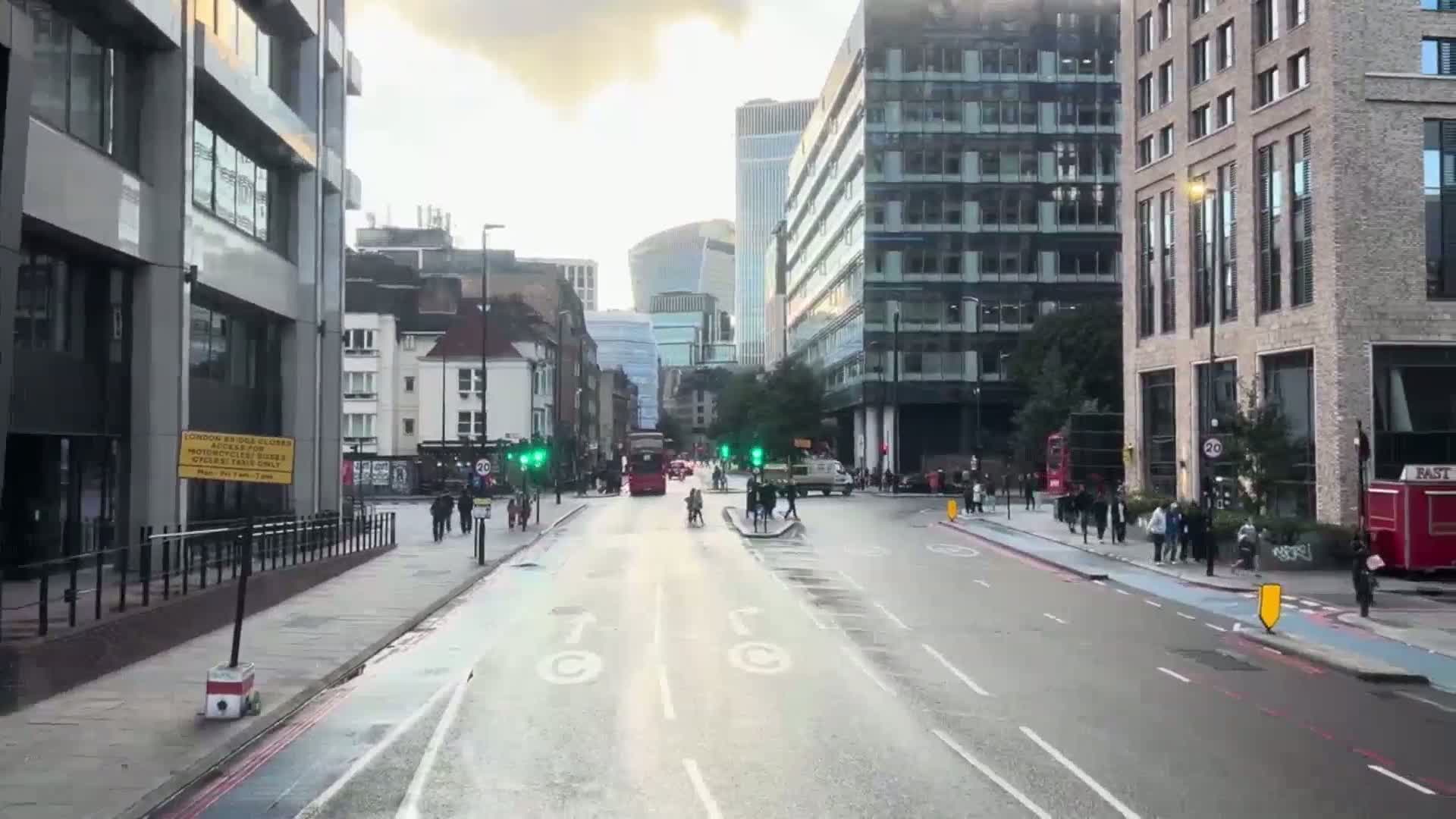 A wet city street scene shows several pedestrians and vehicles, including a double-decker bus, moving past modern and older buildings under a partly cloudy sky.