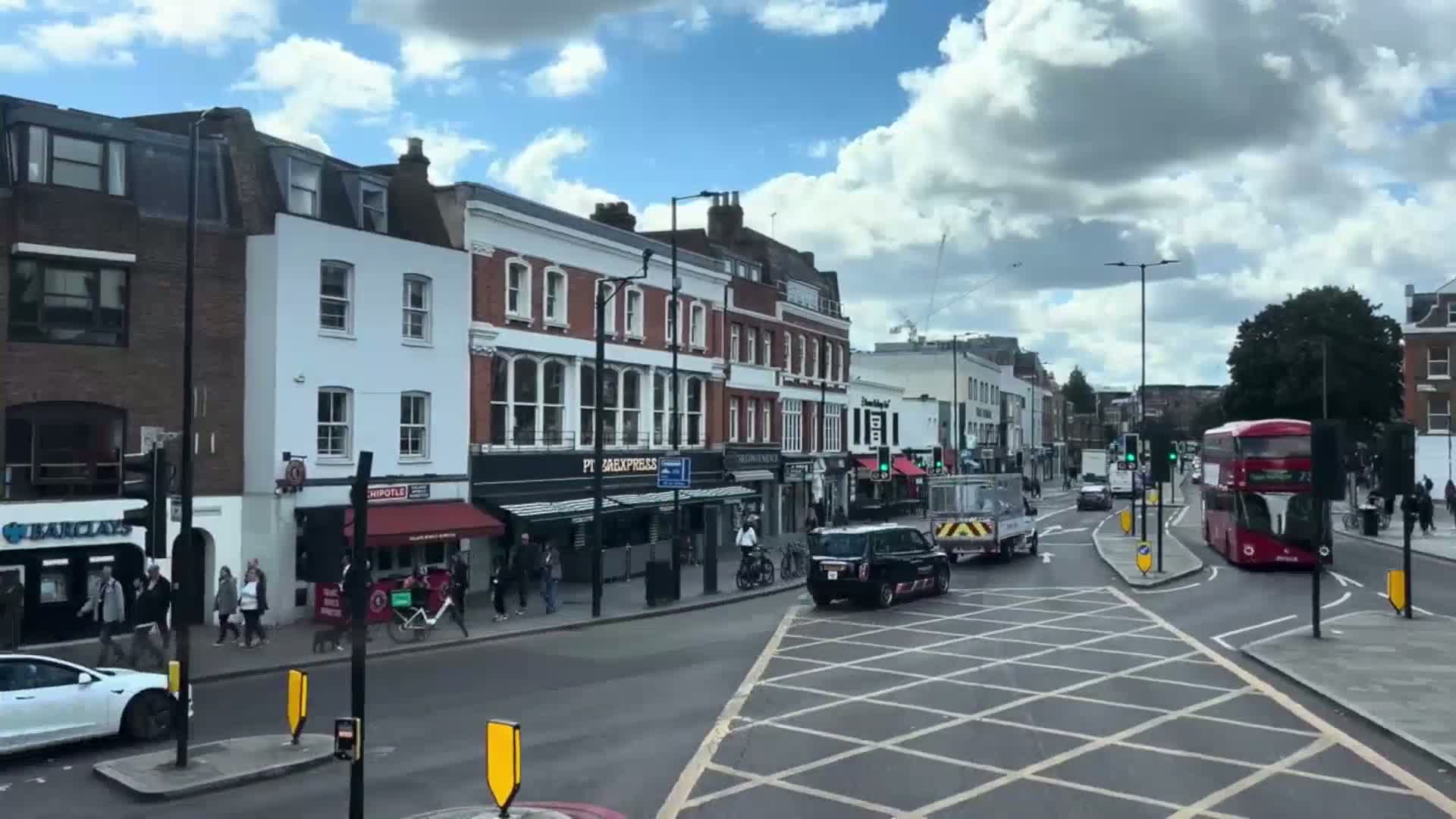 A city street scene under a partly cloudy sky shows pedestrians walking past shops and restaurants, with cars and a double-decker bus navigating a busy intersection.