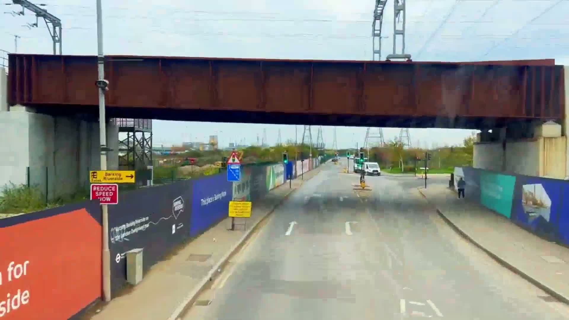 A road passes under a rusty brown railway bridge on an overcast day, with construction barriers and signage lining the sides of the road.