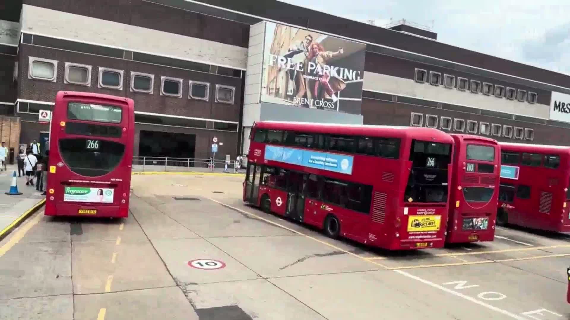 Several red double-decker buses are parked in a lot in front of a large building on an overcast day.