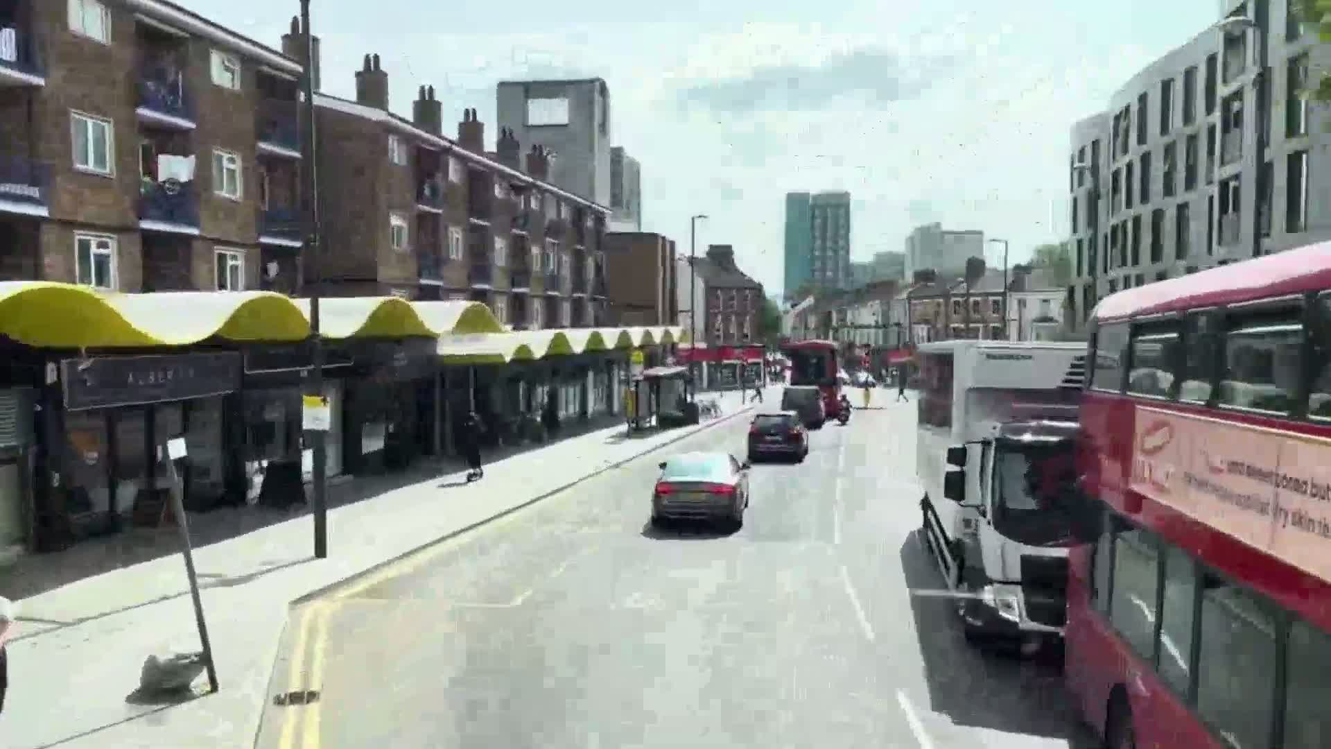 A sunny city street scene shows several cars and buses driving past brick buildings with shops and apartments,  under a mostly clear sky.