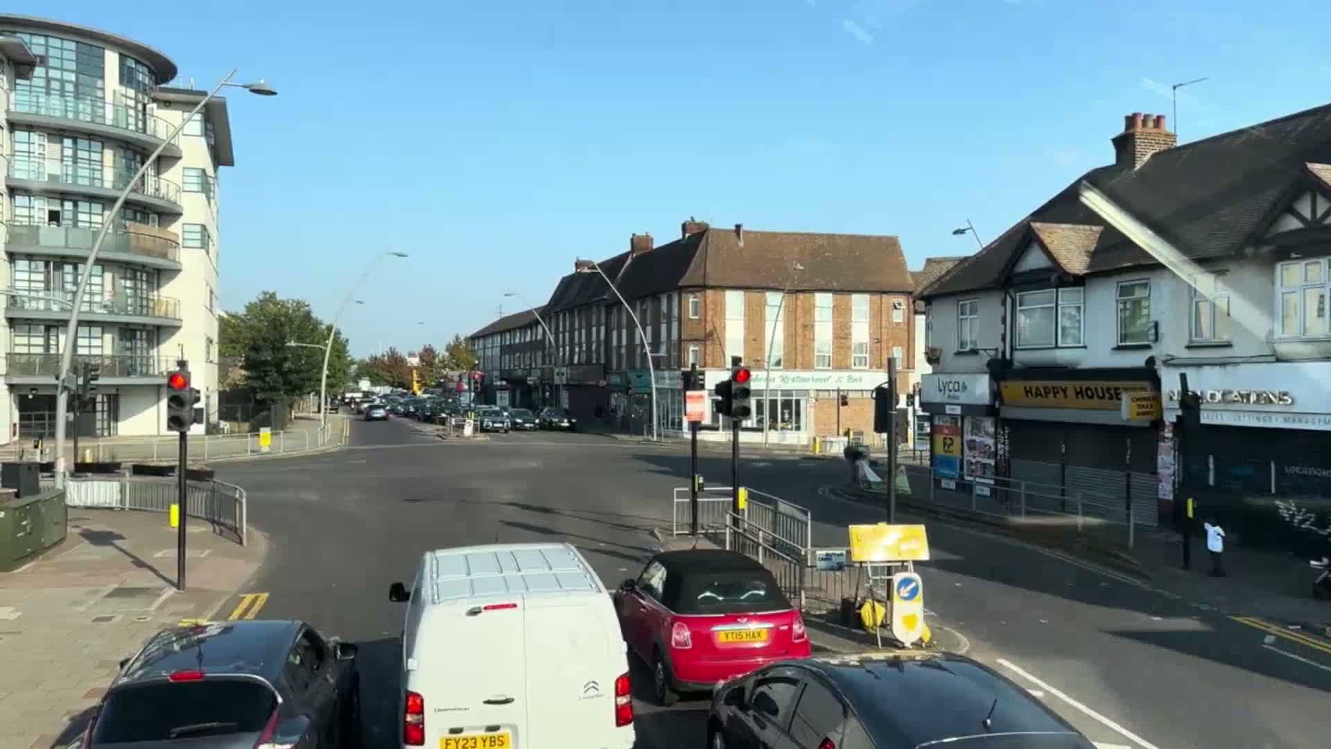 A sunny day shows a street intersection with several cars stopped at a red light, in front of shops and apartment buildings.