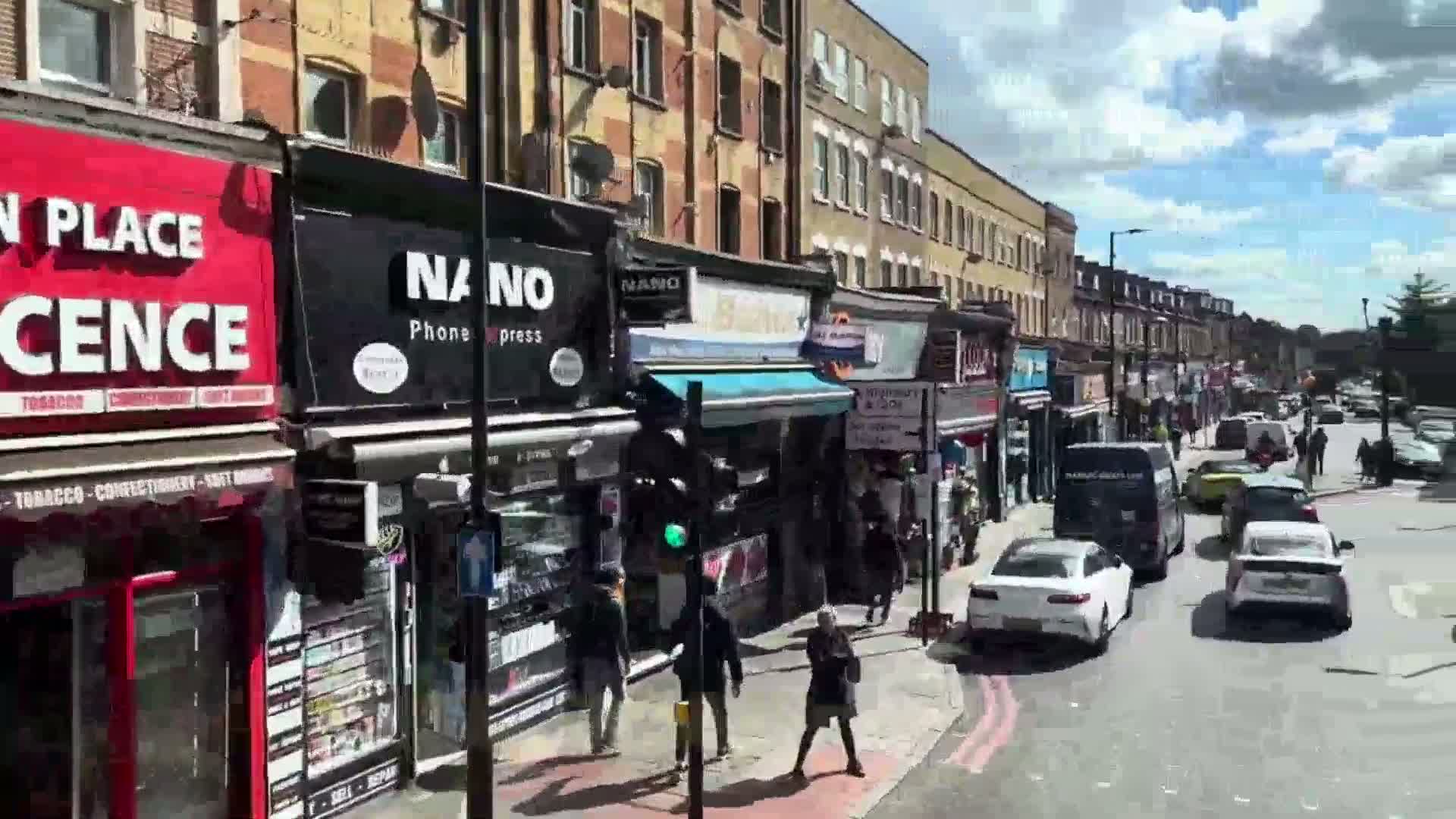 A sunny street scene shows a row of shops with pedestrians and cars on a roadway.
