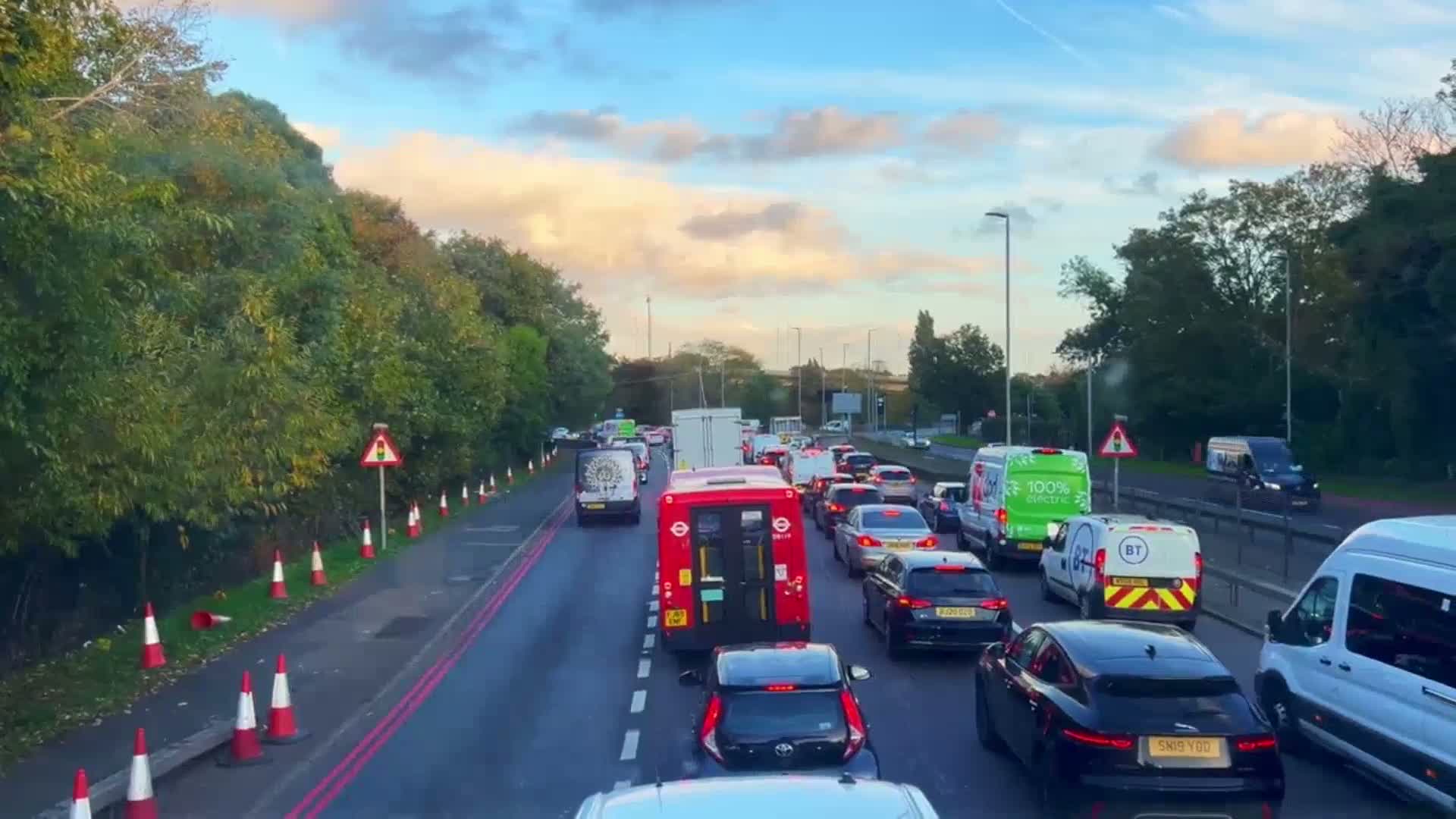 A traffic jam on a multi-lane road under a partly cloudy sky, with green trees lining the roadside and orange traffic cones visible along the shoulder.
