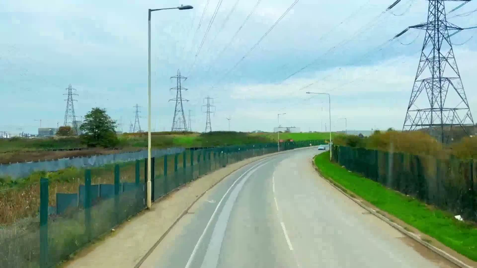 A curved road with a green metal fence on either side, under an overcast sky, passes by several tall electricity pylons and some low-lying vegetation.
