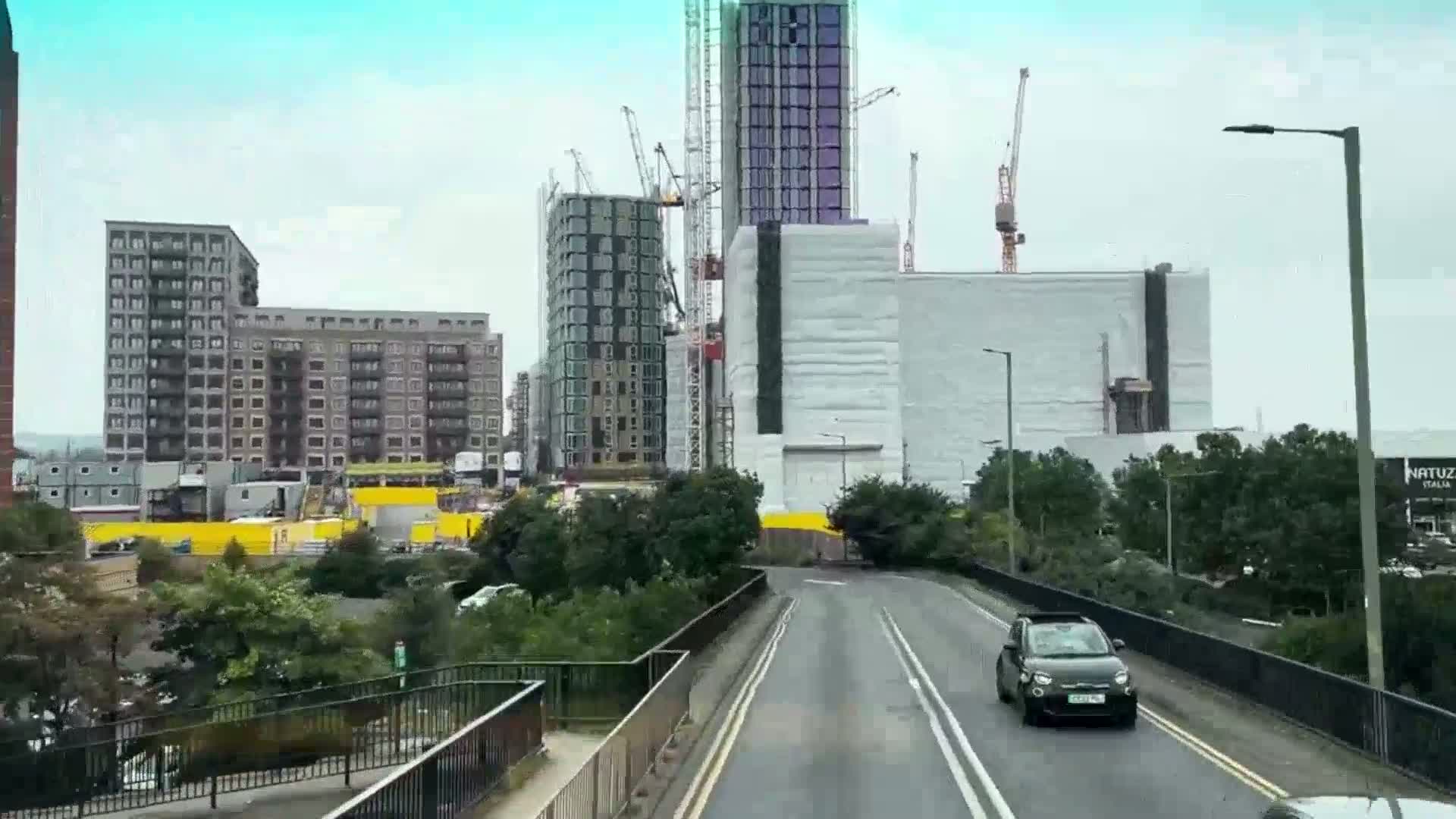A road passes under a bridge, flanked by green vegetation and numerous high-rise buildings under construction, some partially shrouded in white sheeting, in overcast weather.