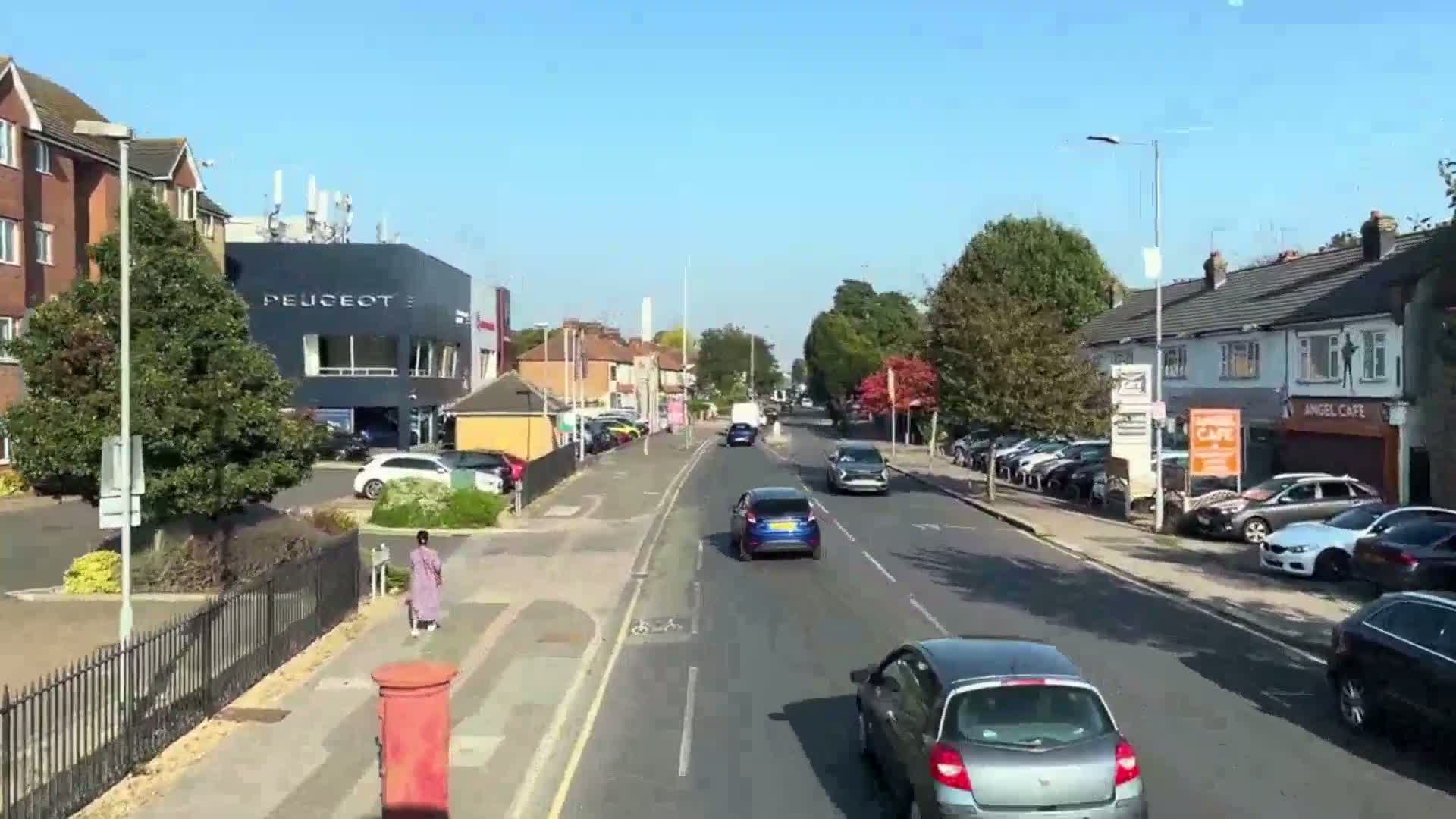 A sunny day shows a residential street with cars parked on both sides, a few cars driving, and a person walking along the sidewalk, passing a postbox.