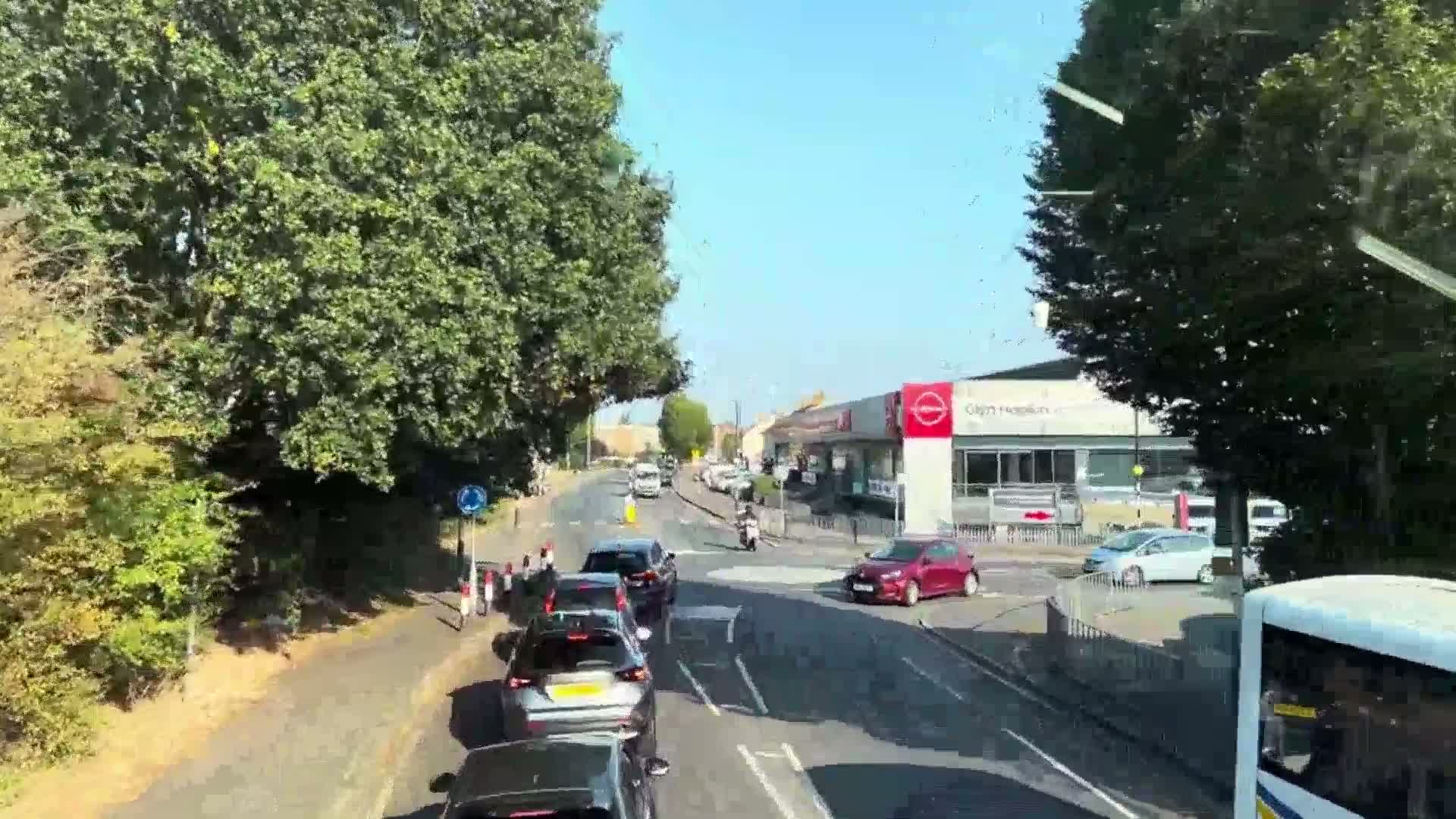 A sunny day reveals a line of cars stopped at a road intersection, with a motorcyclist and pedestrians visible nearby, all surrounded by lush green trees and buildings.
