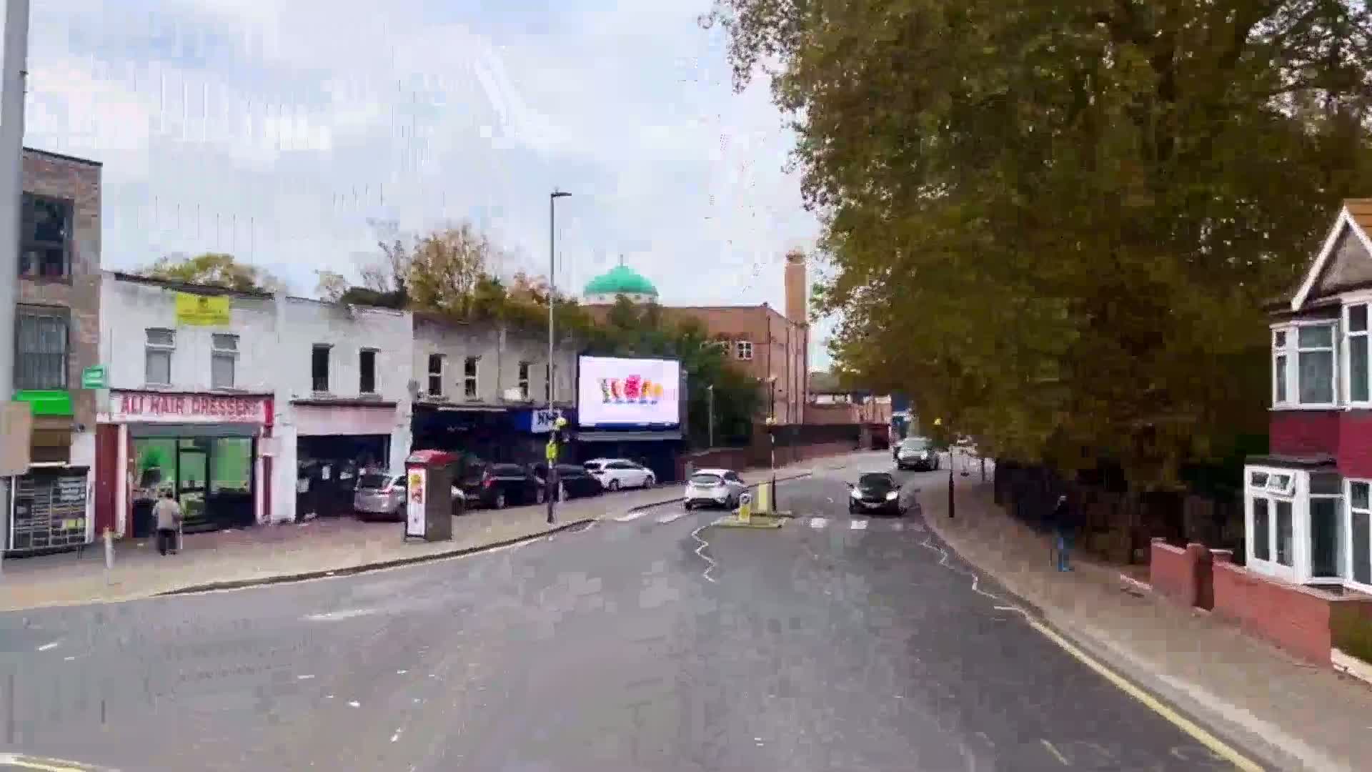 A street scene on an overcast day shows several cars at an intersection near shops and a mosque with a large digital billboard.