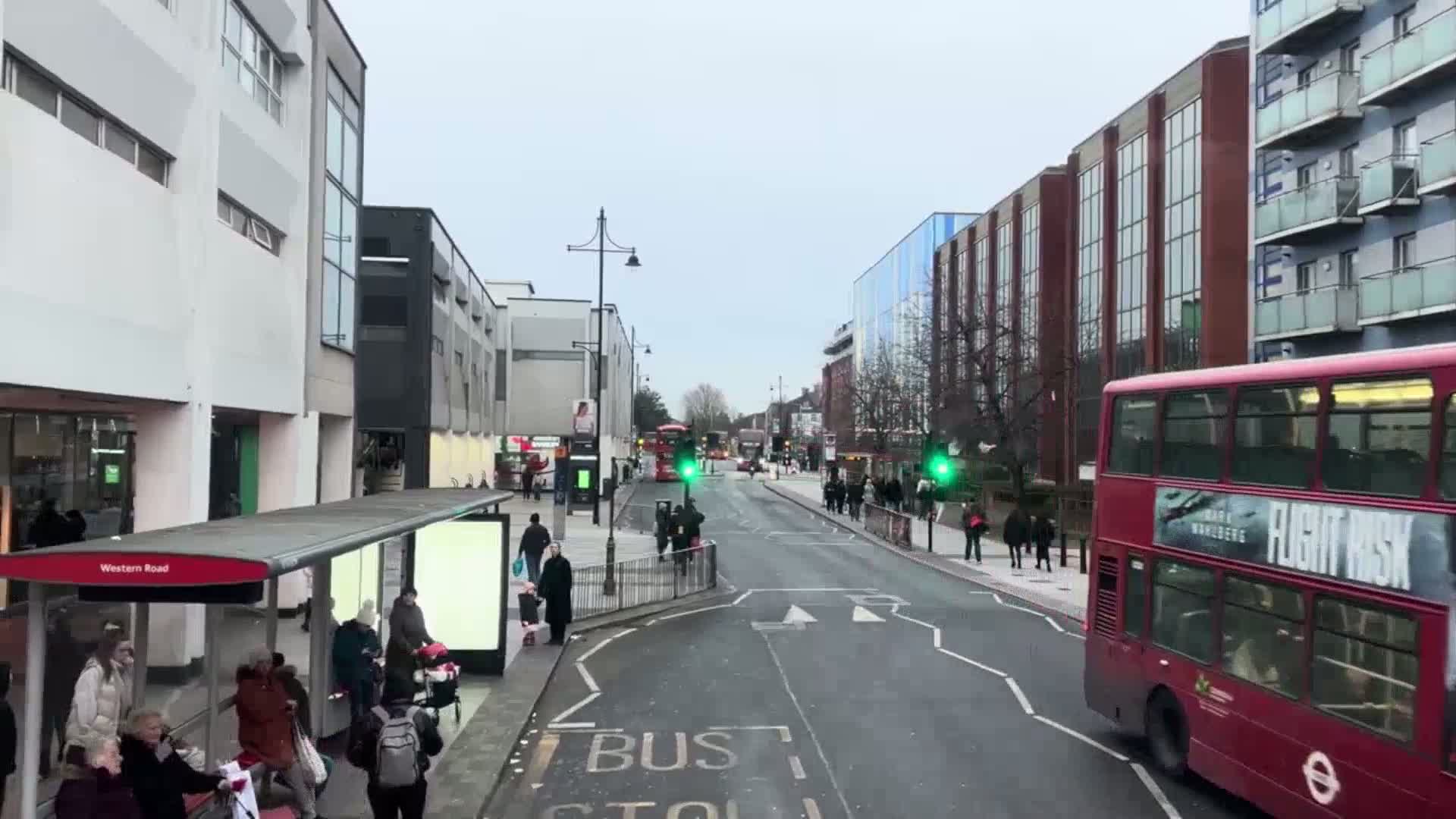 A city street scene on an overcast day shows pedestrians waiting at a bus stop and several buses on a street lined with multi-story buildings.