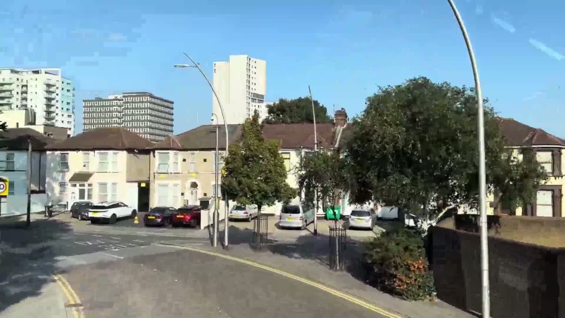 A sunny day reveals a residential street scene with two-story houses, parked cars, and taller buildings in the background.