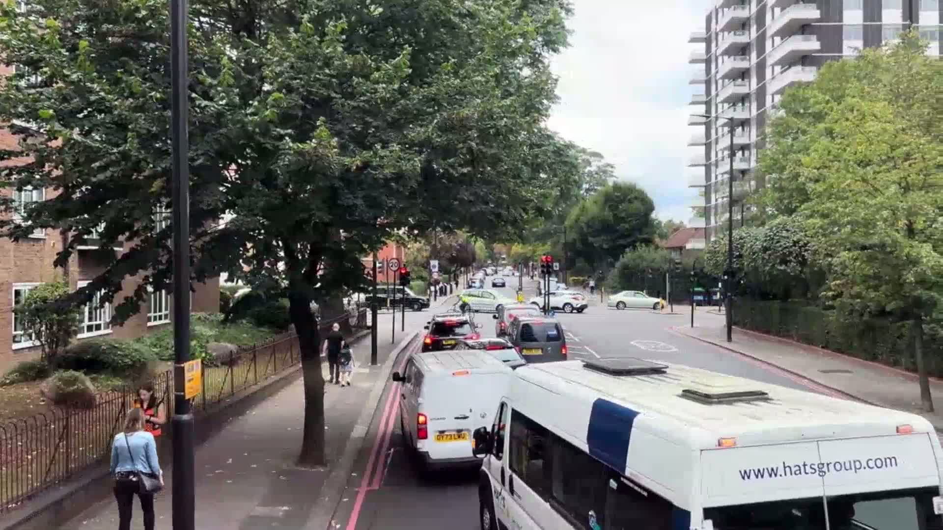 A view from a bus shows a moderately congested street with pedestrians on the sidewalk under an overcast sky, lined with buildings and trees.