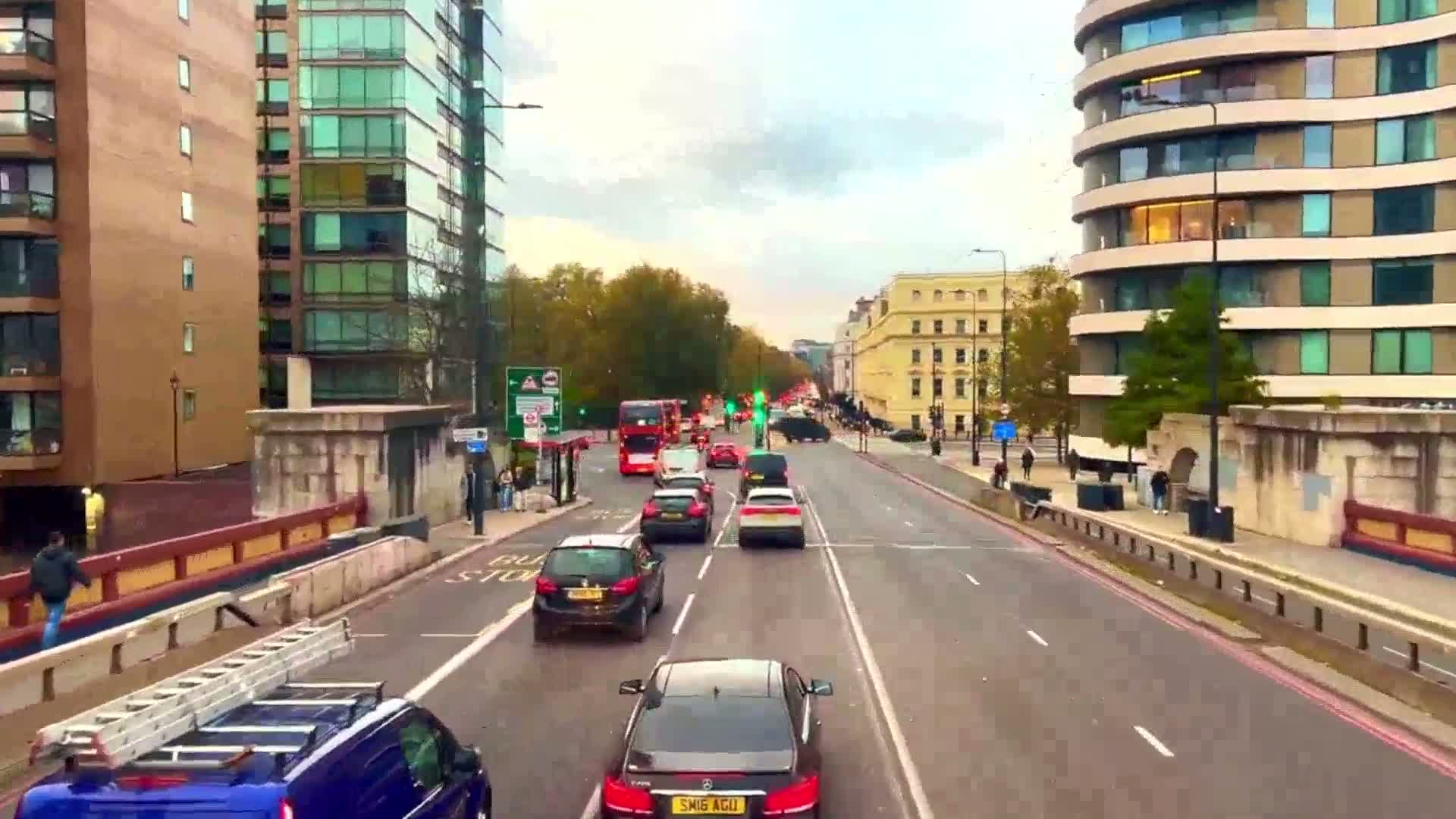 A city street with several cars and buses driving in both directions under a partly cloudy sky, lined with buildings on either side.