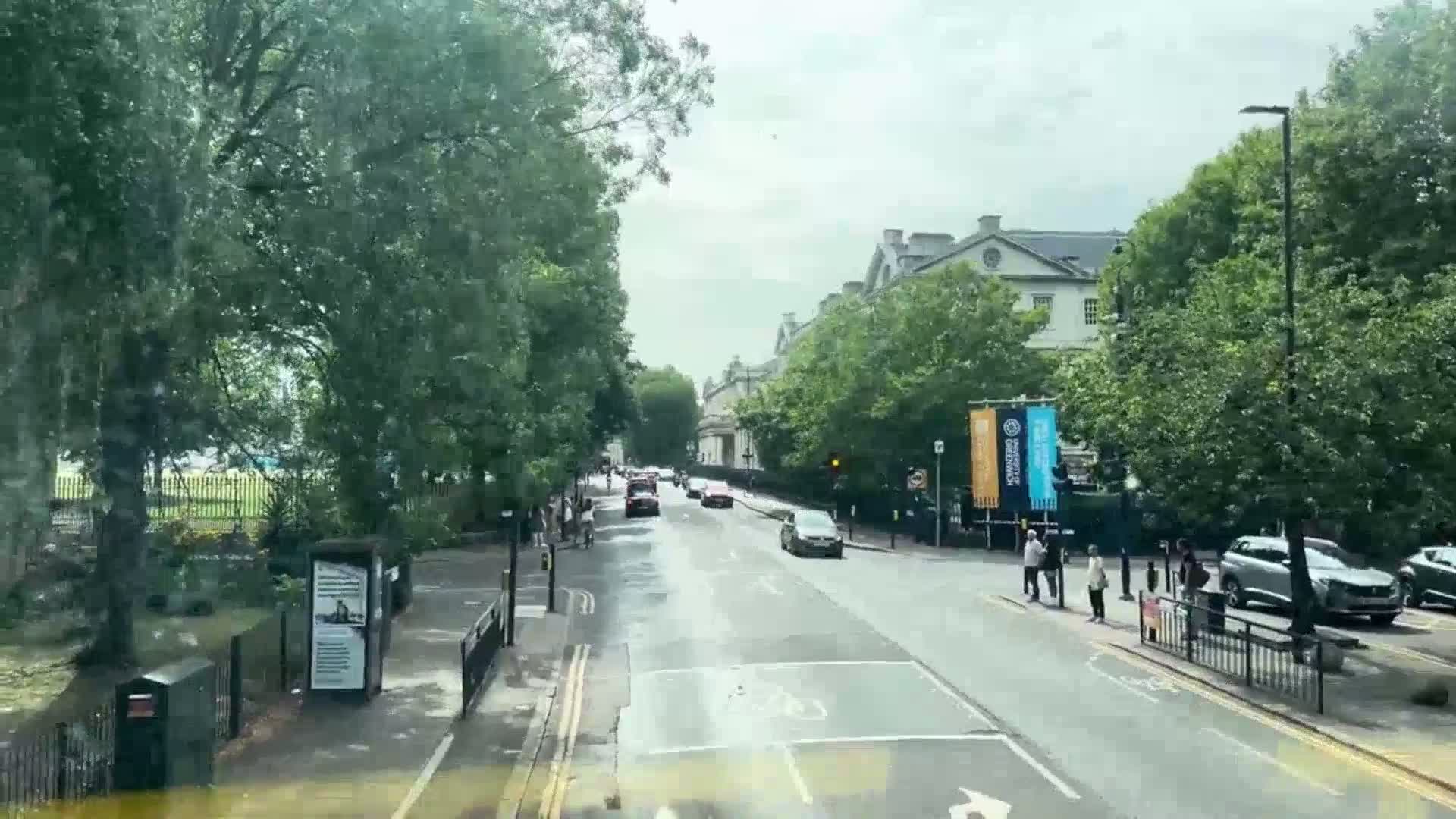 A wet street lined with trees and buildings, with several cars and pedestrians visible.