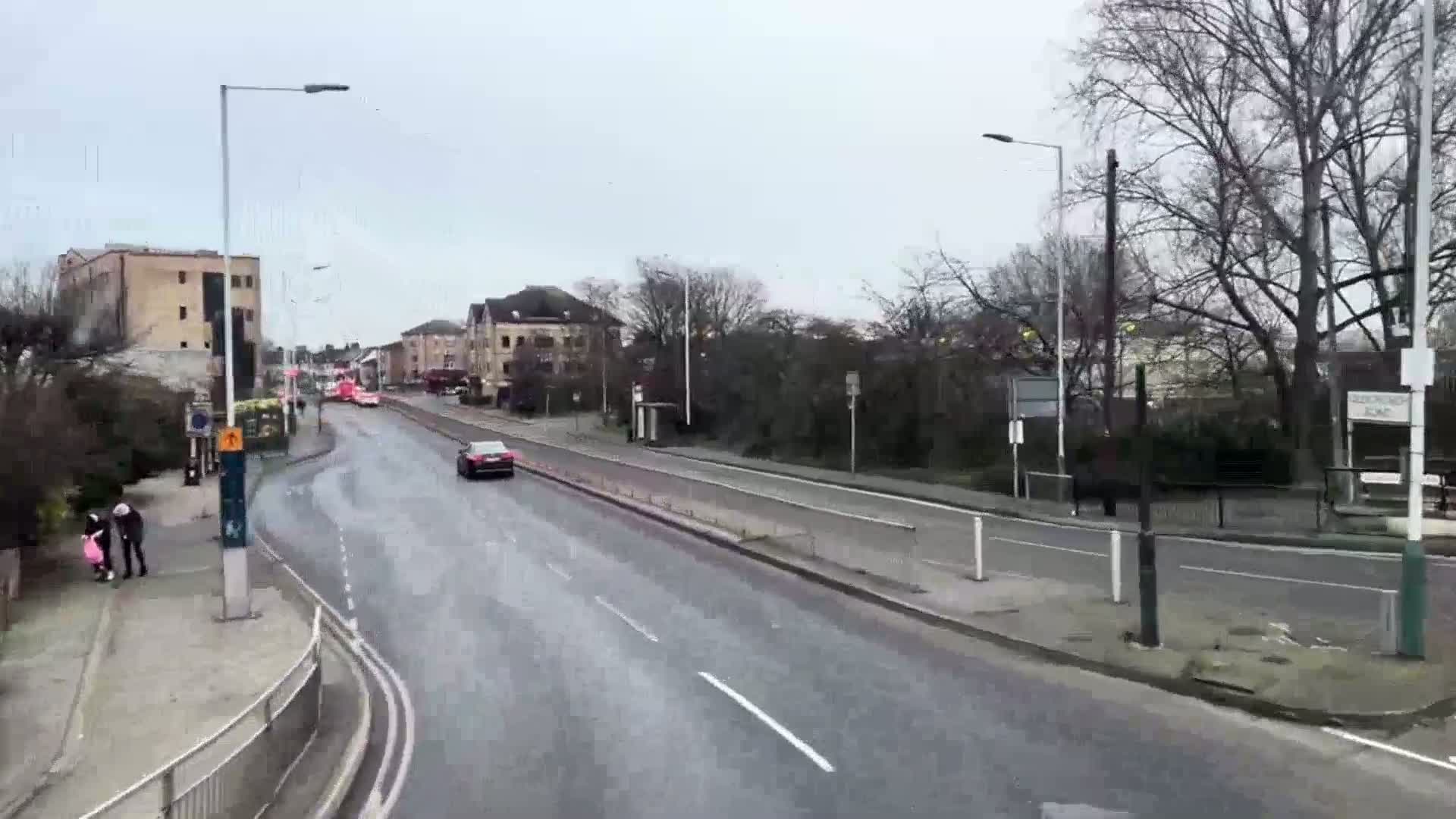 A wet, overcast day shows a multi-lane road with sparse traffic, buildings lining the road, and a few pedestrians on the sidewalk.