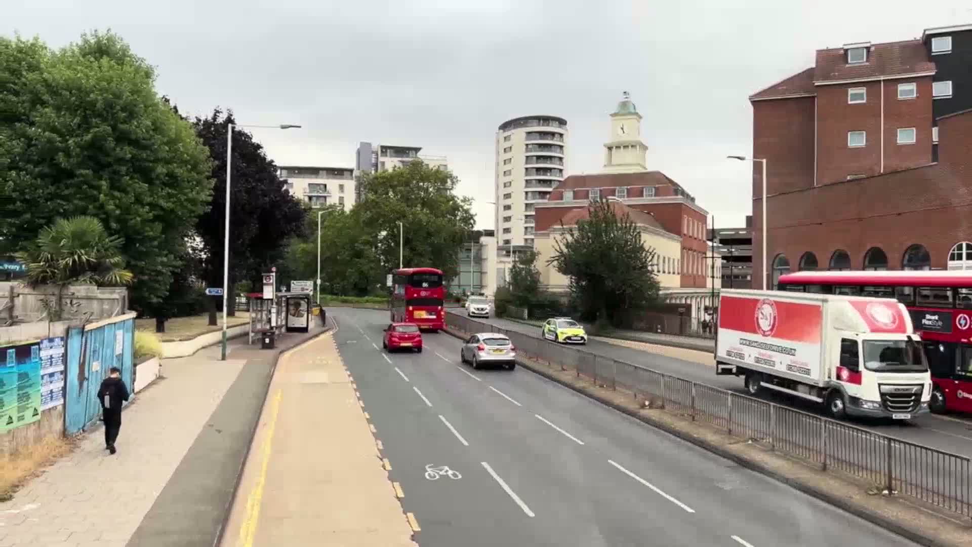 A city street scene on an overcast day shows several vehicles including two double-decker buses, cars, and a delivery truck, traveling along a road with a bus stop and pedestrian walking alongside.