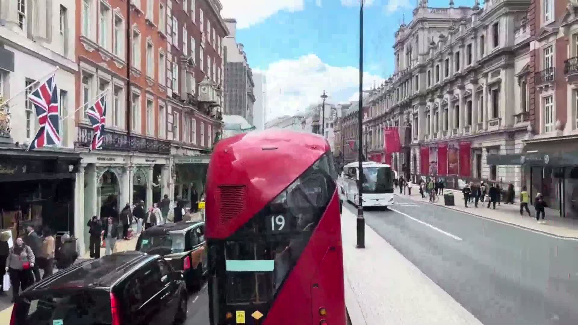 A red double-decker bus drives down a city street on a sunny day past pedestrians and other vehicles, with tall buildings lining both sides of the road.