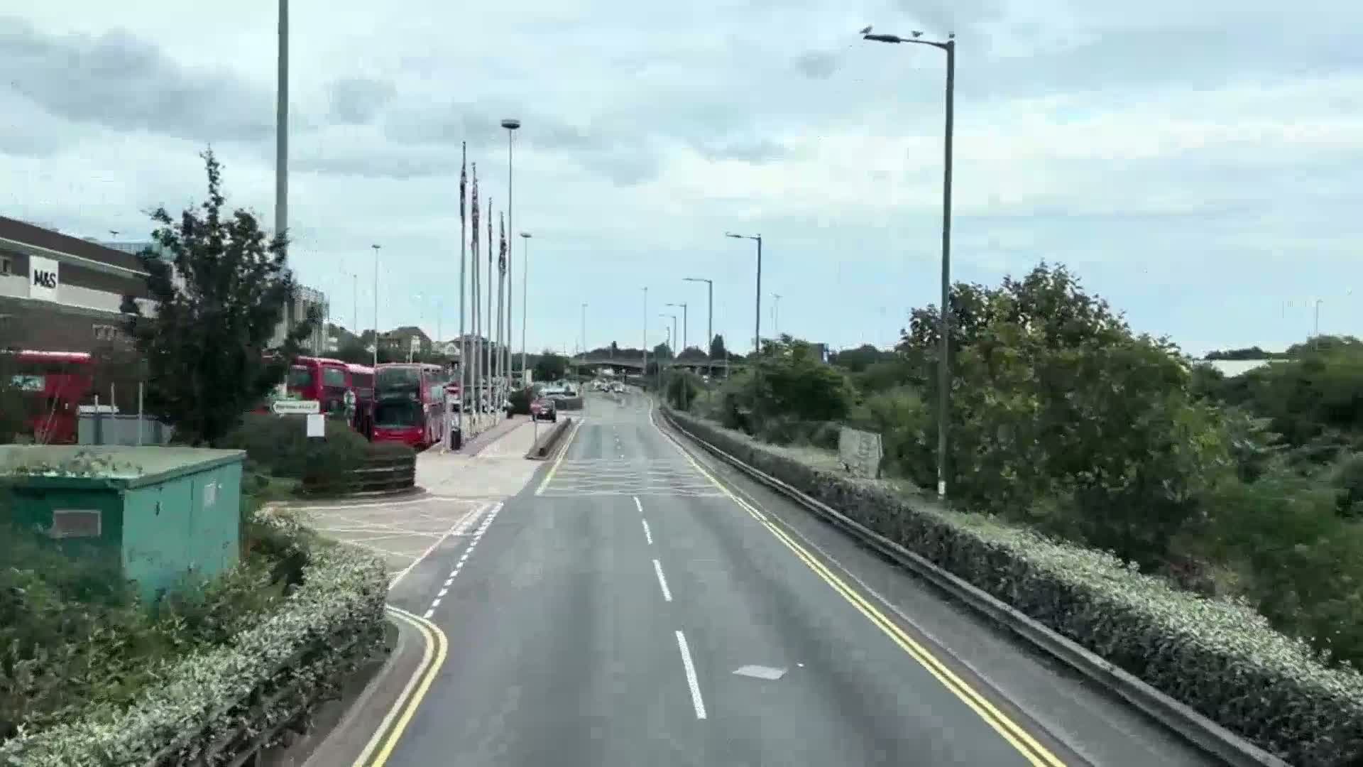 A multi-lane road stretches ahead under an overcast sky, with several red double-decker buses parked at a bus stop near a building to the left.