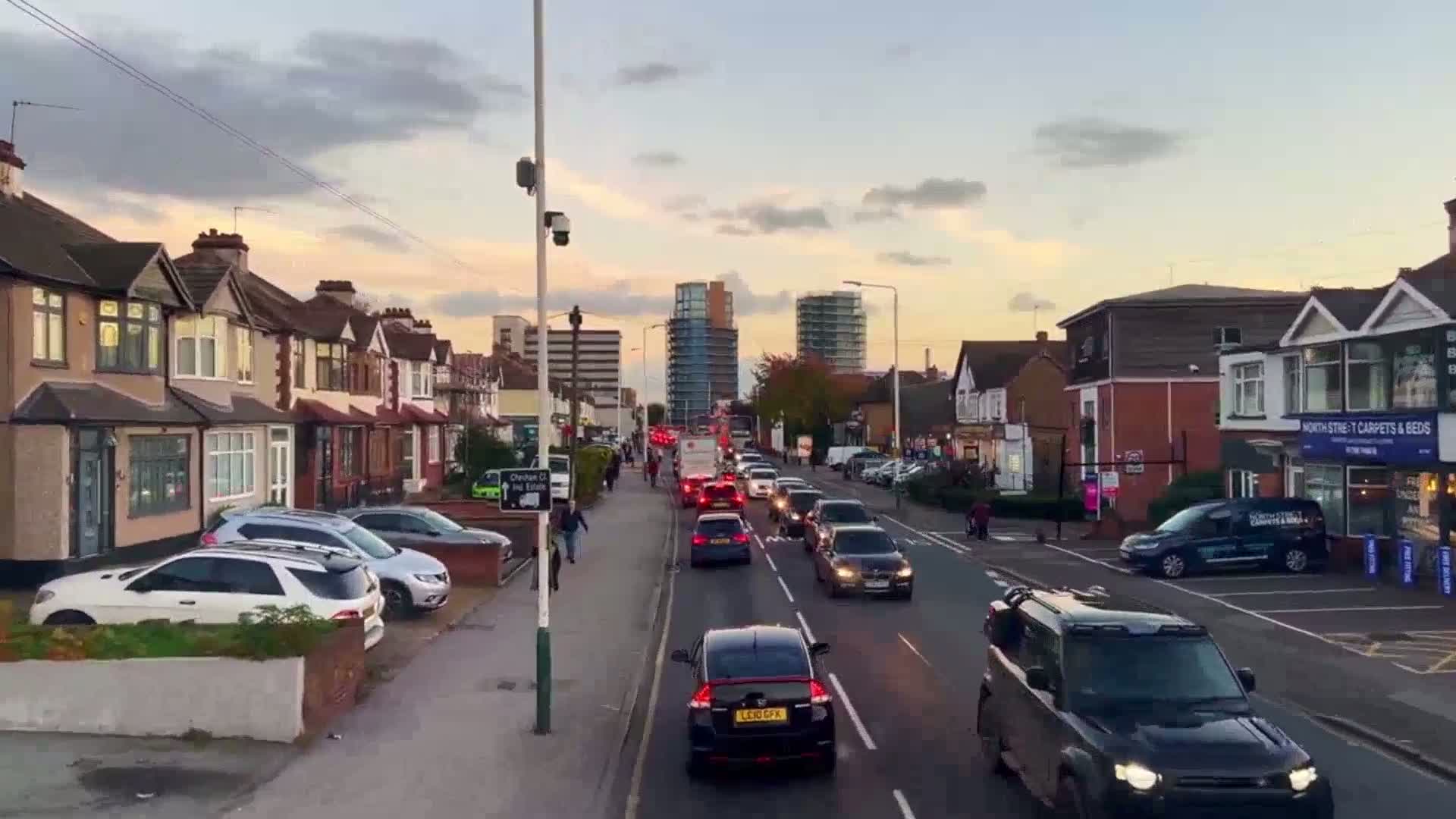 Under a partly cloudy, dusky sky, a street lined with residential houses and shops shows moderate traffic congestion.