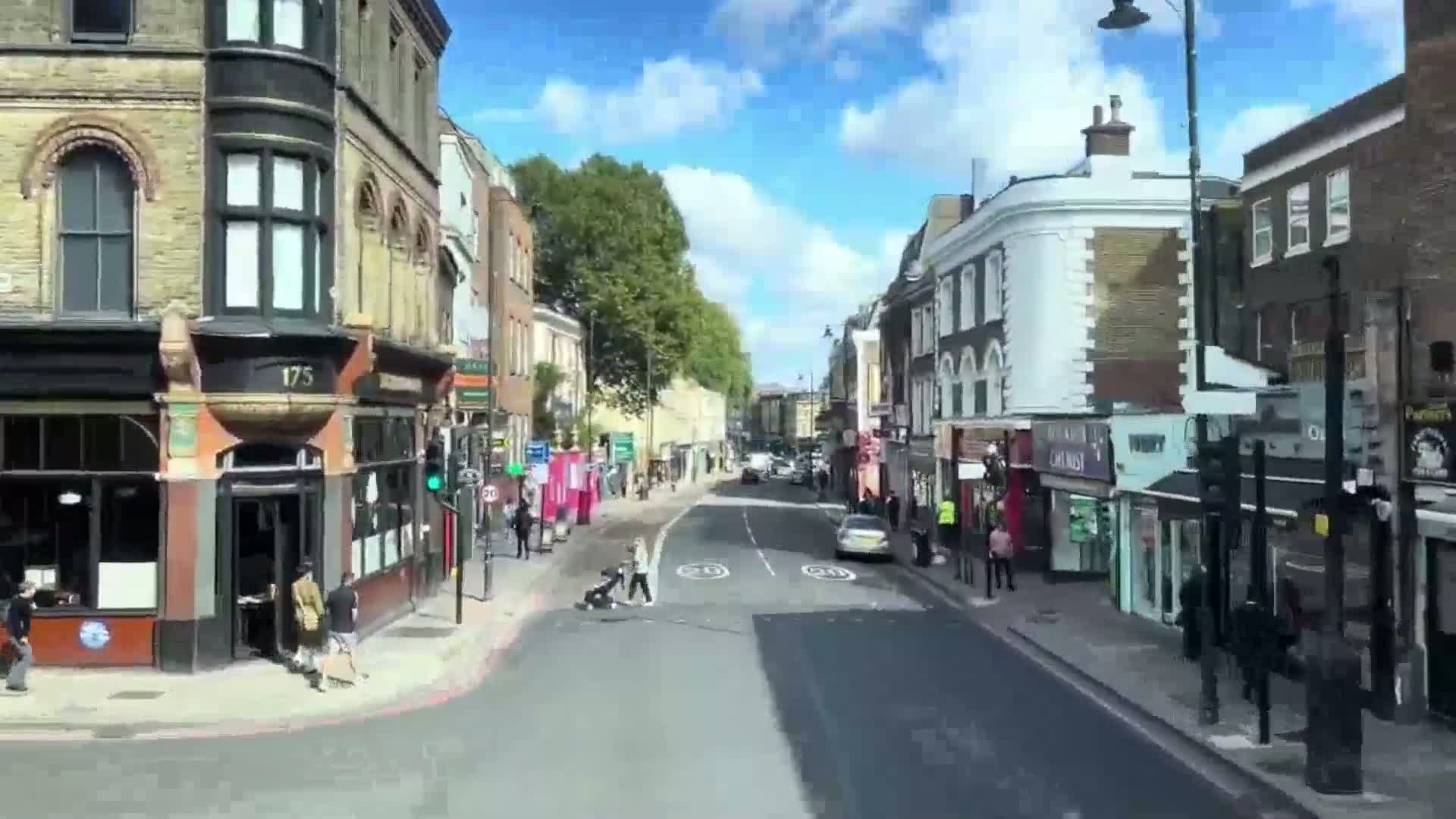 A sunny day on a city street lined with multi-story brick buildings and shops, with several pedestrians walking on the sidewalk and crossing the street.