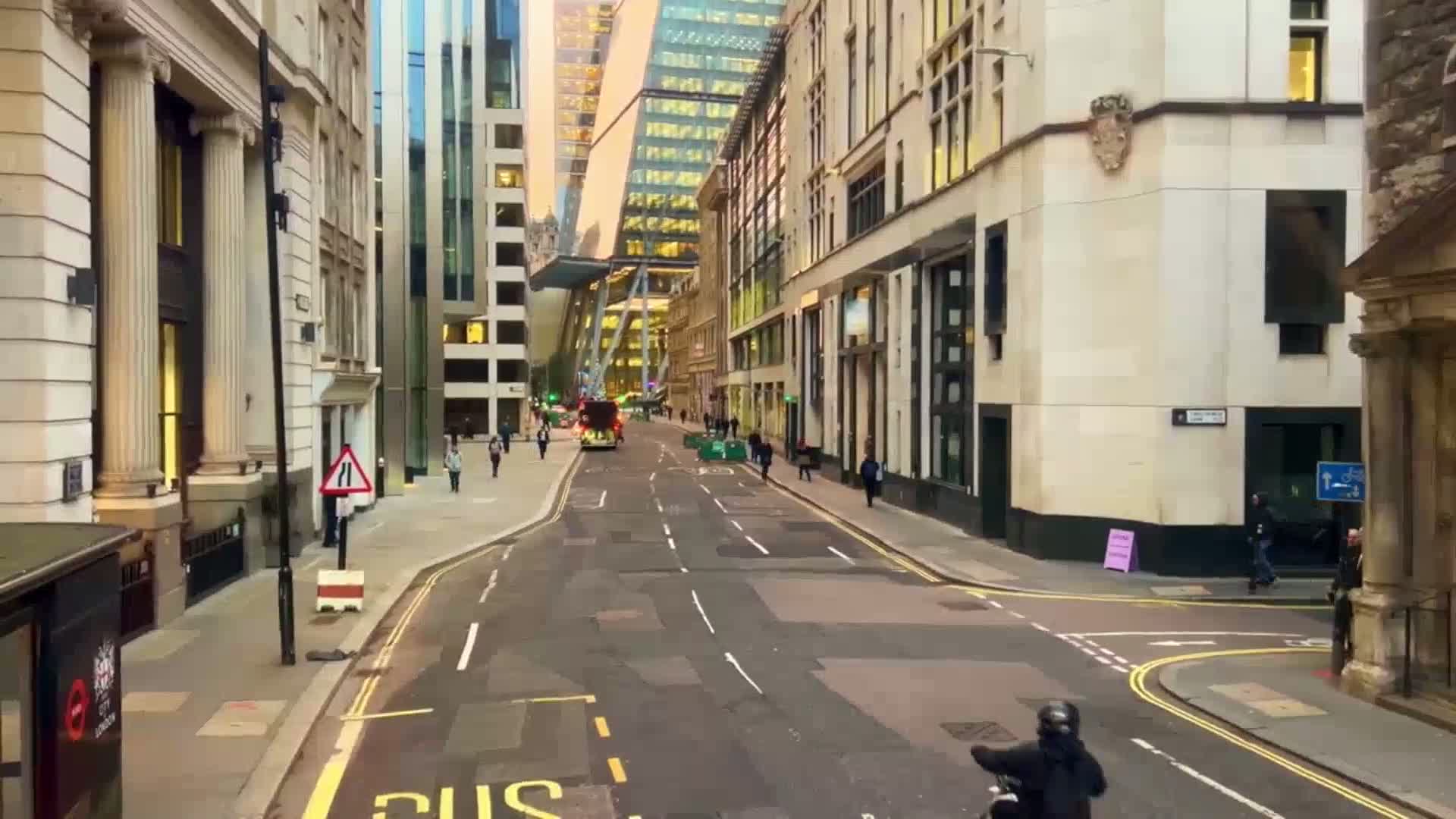 A city street lined with tall buildings, under a partly cloudy sky, with a few pedestrians and a single vehicle on the road.
