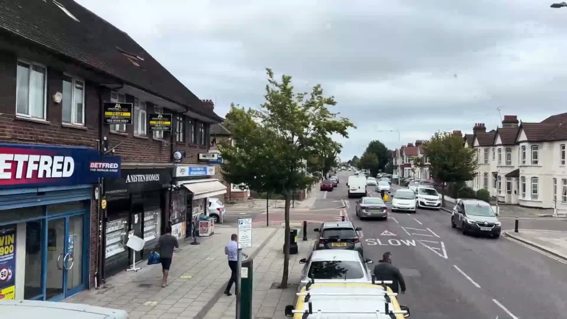 A street scene on an overcast day shows several cars driving past shops and residential houses.