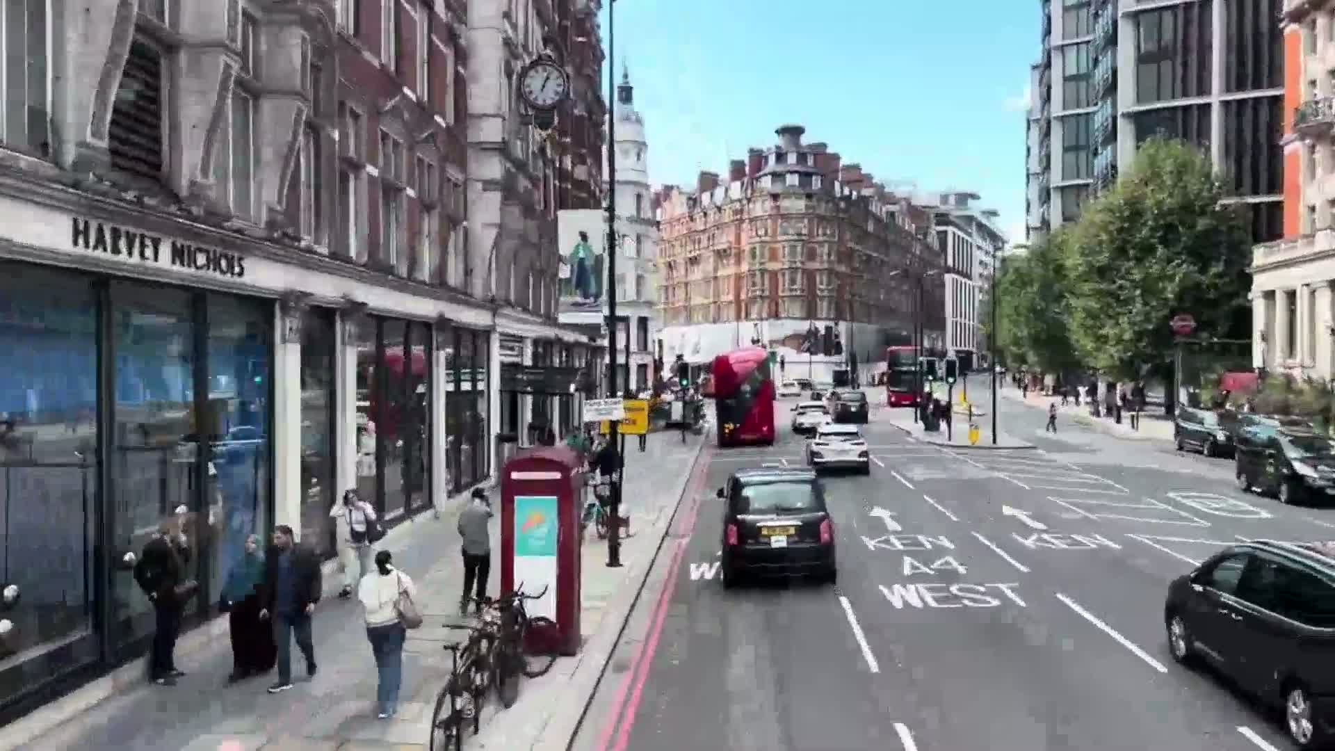A sunny city street scene shows several pedestrians, parked bicycles, and various vehicles including double-decker buses and cars, along the sides of multi-story buildings.
