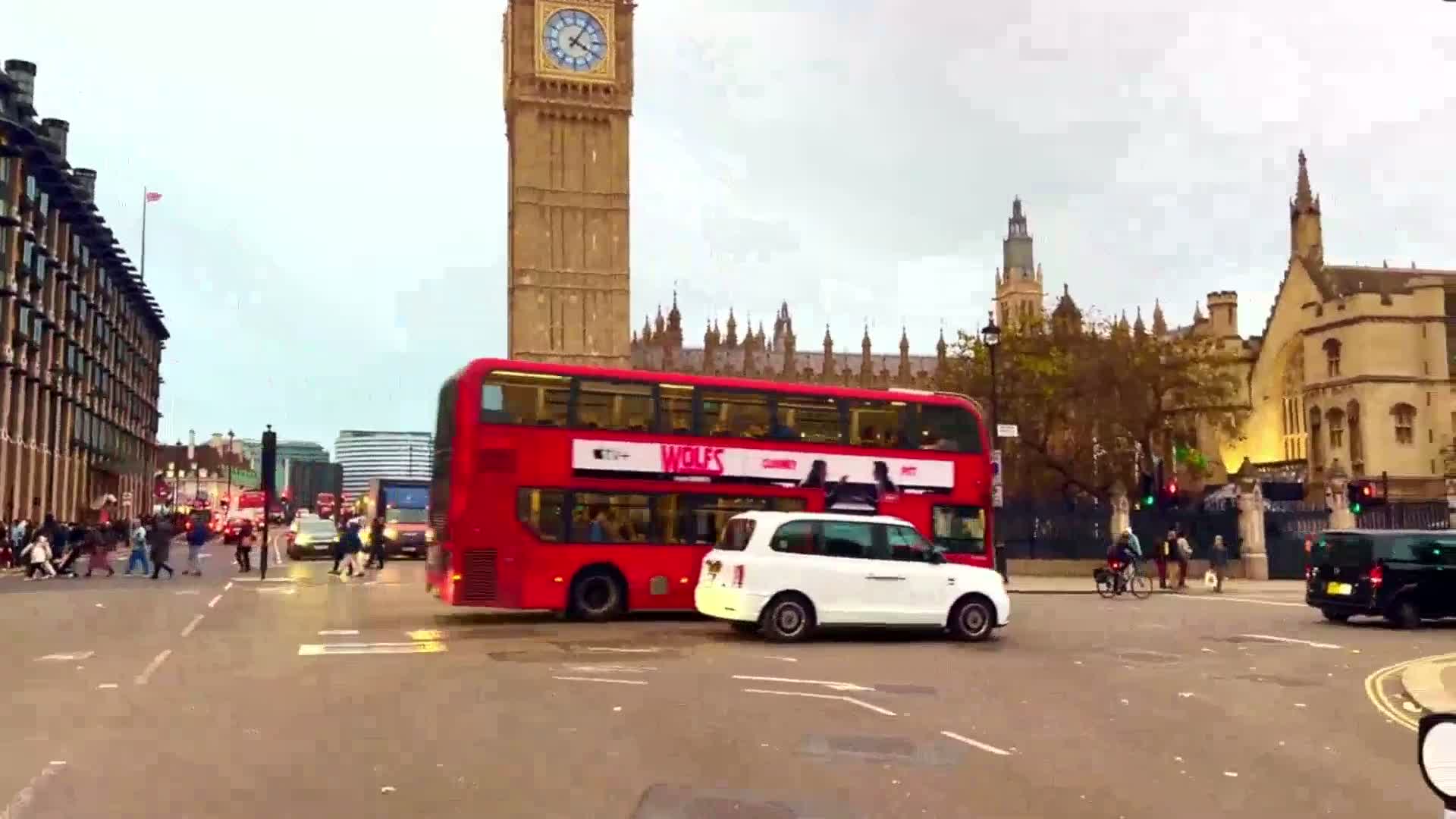 A red double-decker bus and a white taxi are in a London street intersection near Big Ben, with pedestrians crossing and overcast weather conditions.