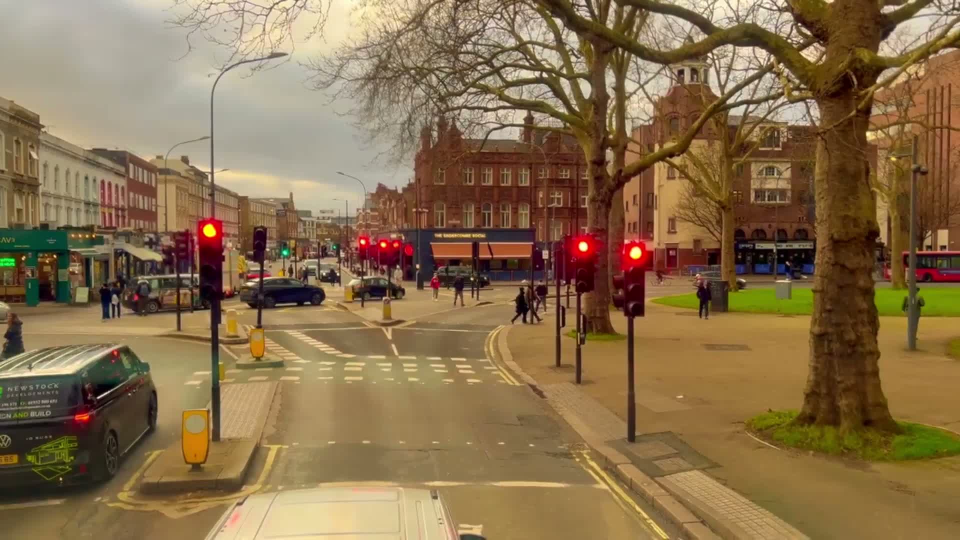 A street scene shows overcast skies, leafless trees, traffic stopped at red lights, pedestrians crossing, and cars and buses driving along a multi-lane road in front of brick buildings and shops.