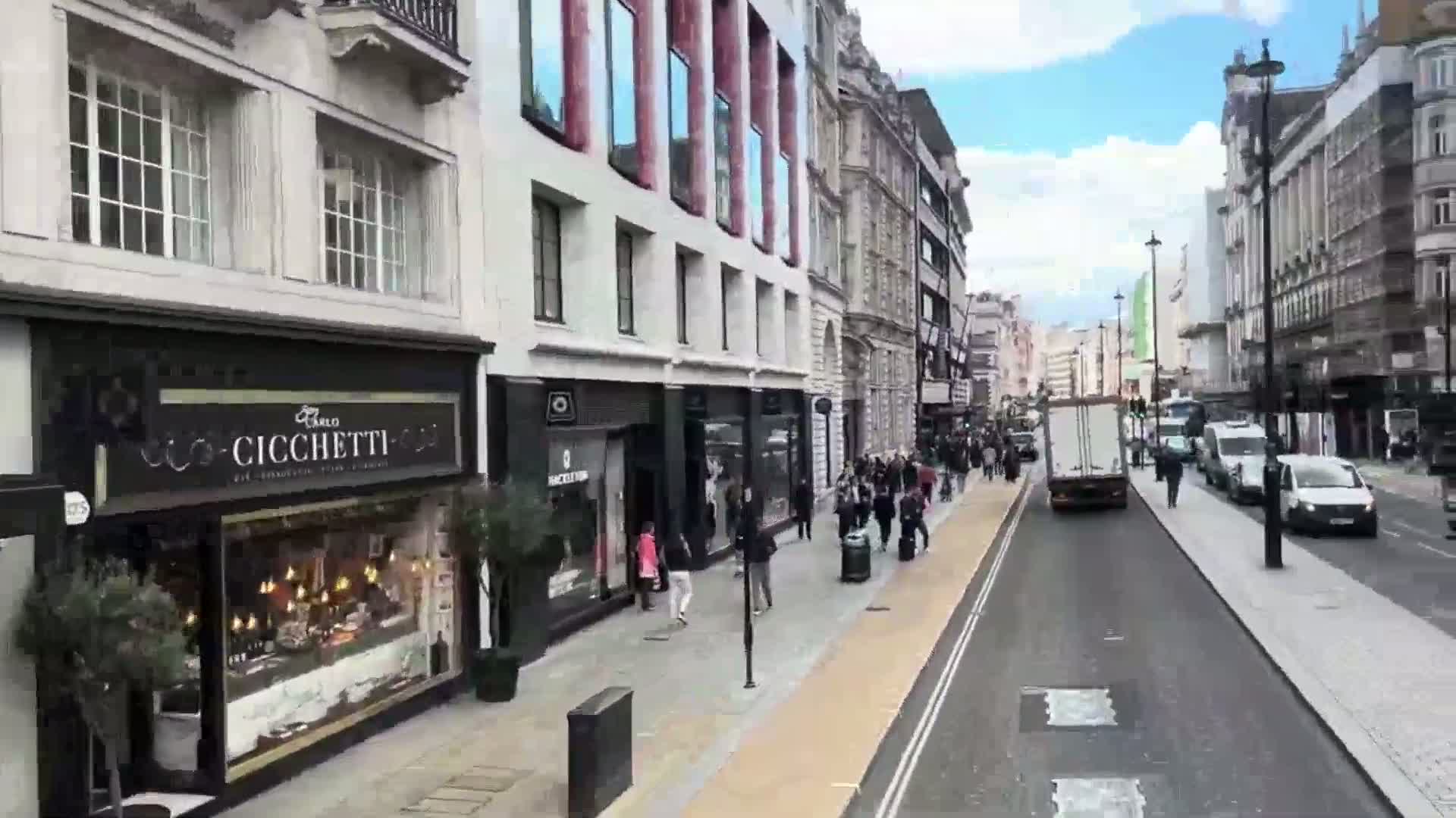 A city street scene on a partly cloudy day shows pedestrians walking past shops and businesses, with several vehicles driving on the road.