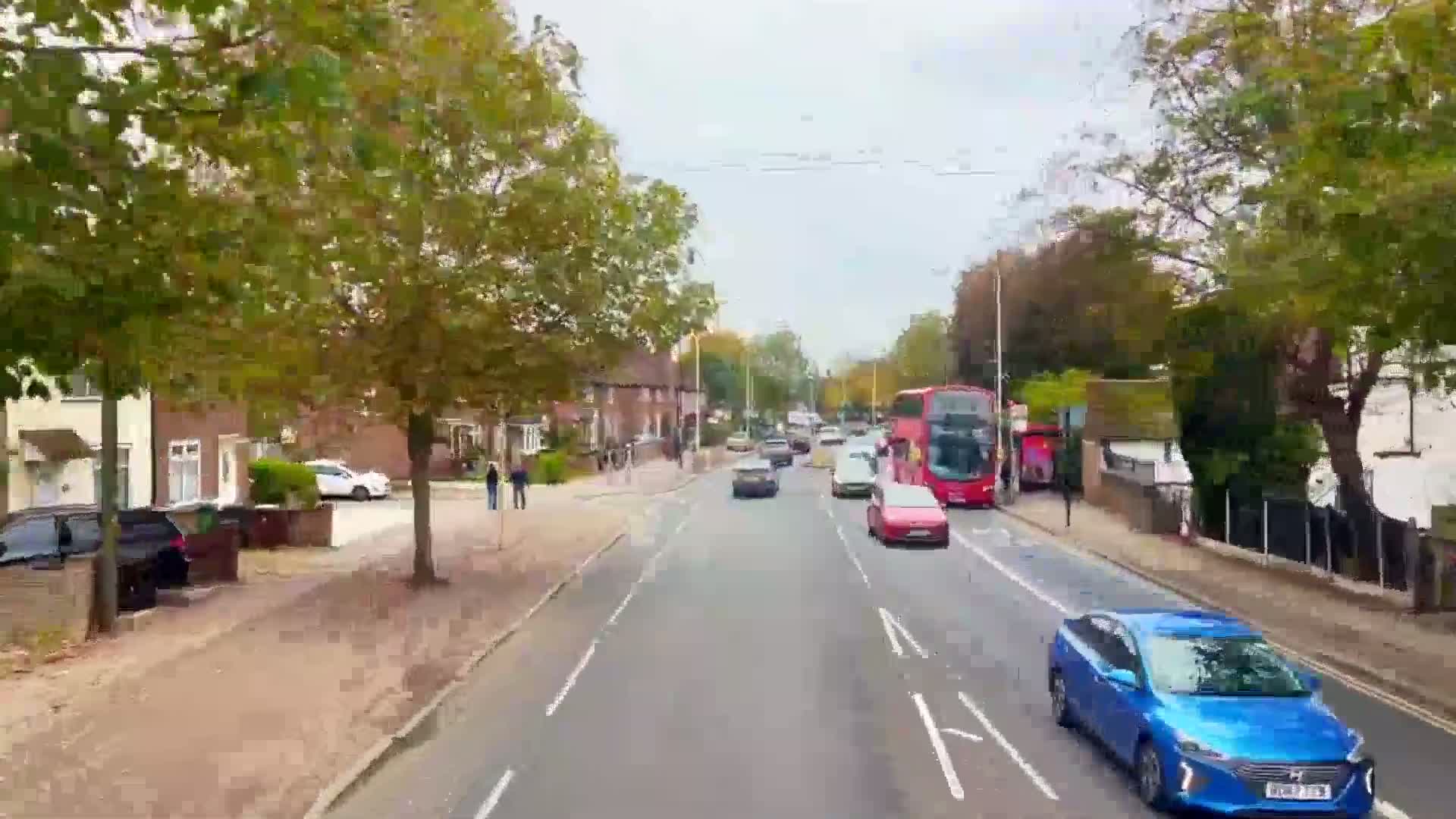 A street scene on an overcast day shows several cars driving on a two-lane road lined with houses and trees, with a double-decker bus visible in the distance.
