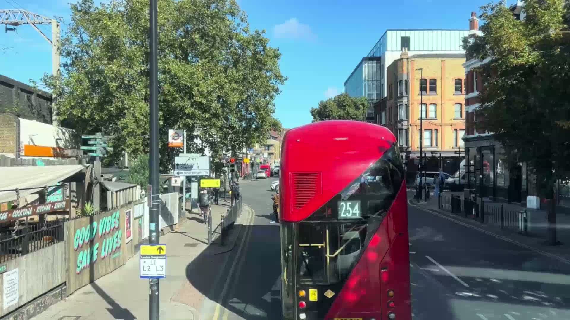 A red double-decker bus drives down a sunny street lined with buildings and trees, past pedestrians and other vehicles.