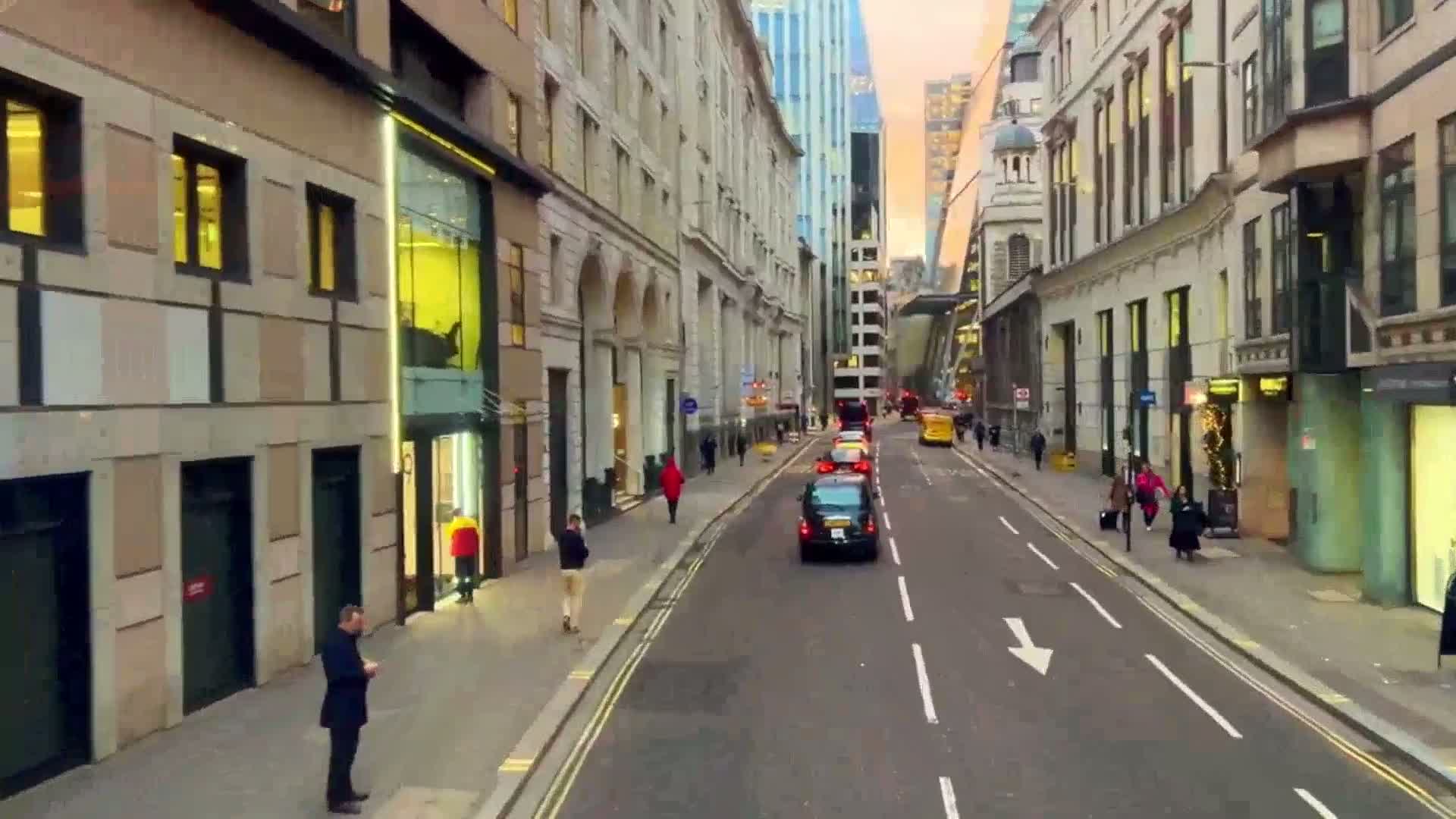 A city street lined with tall buildings, some pedestrians, and several cars driving in both directions under a dusky sky.