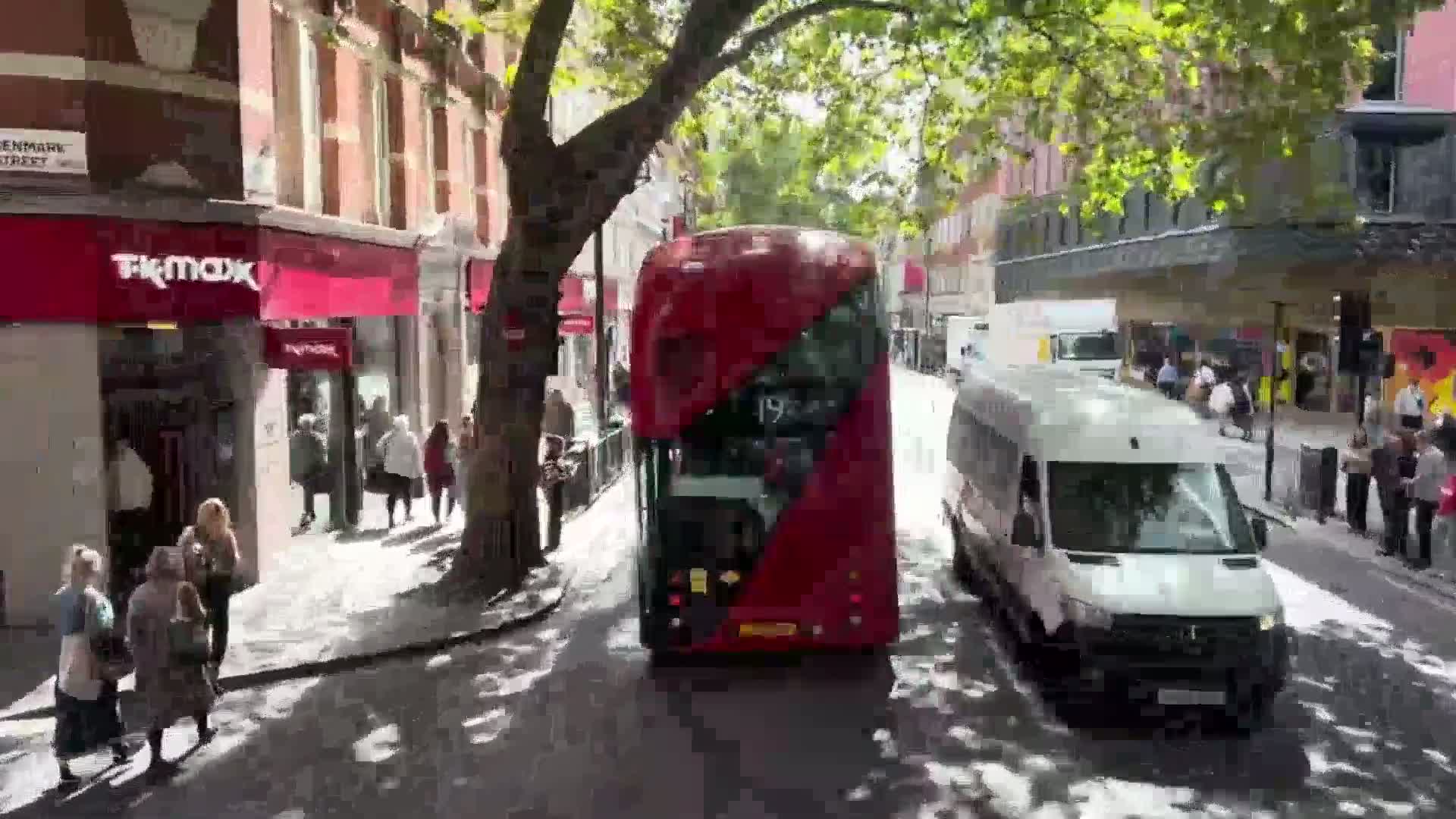 A double-decker bus and a white van are driving down a city street on a sunny day, past pedestrians and shops.