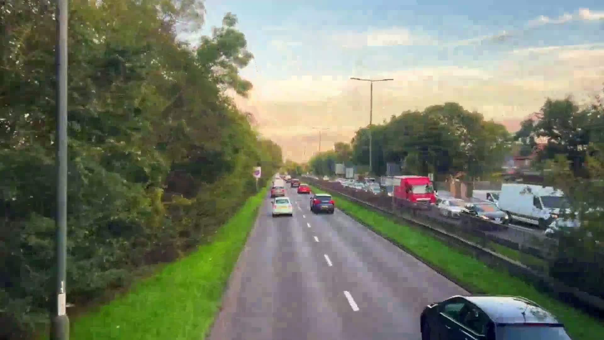 A multi-lane road with moderate traffic flows under a partly cloudy, sunset sky, lined with green vegetation.