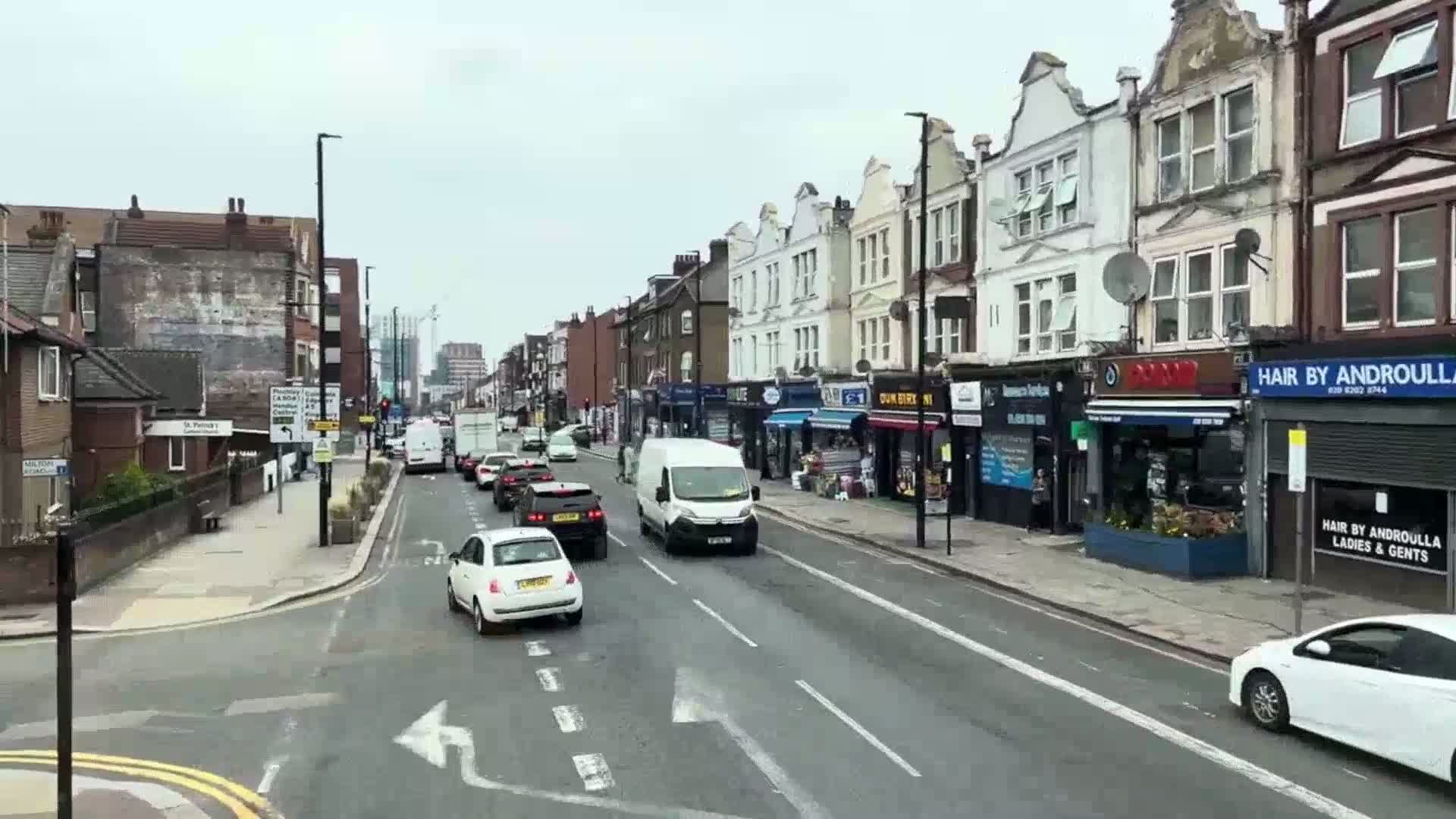 A street lined with two-story shops and residential buildings is moderately trafficked with cars under an overcast sky.