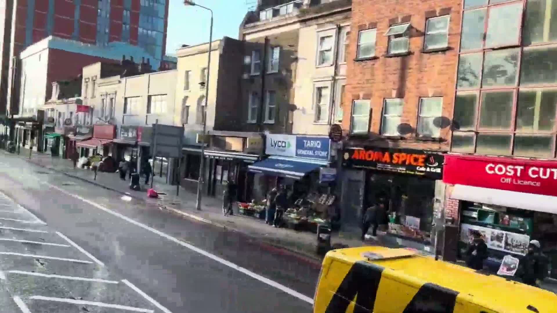 A wet city street scene on a partly sunny day shows multiple shops and pedestrians in front of brick buildings.