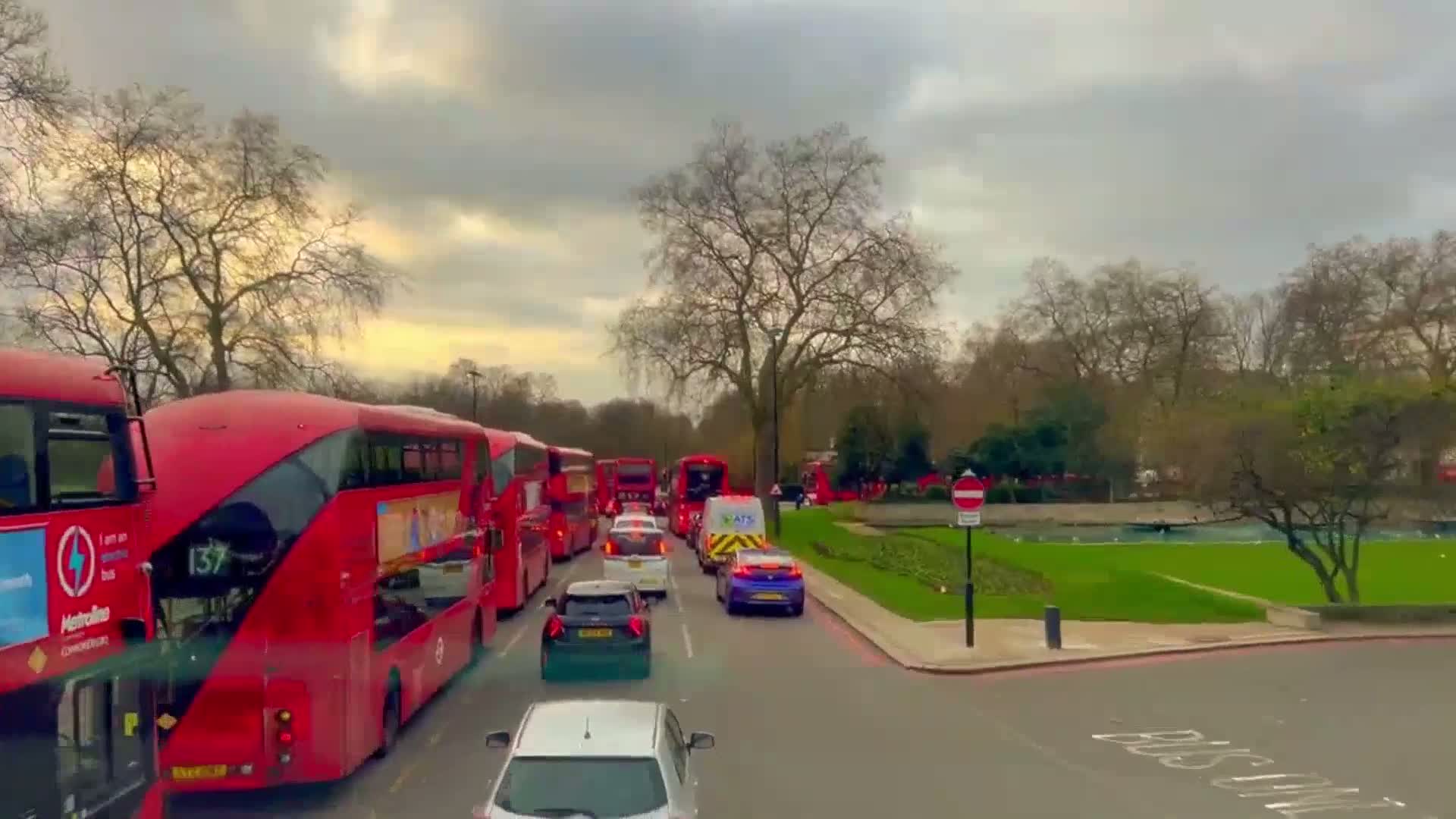 A line of red double-decker buses sits in traffic on a street next to a park under a cloudy sky.