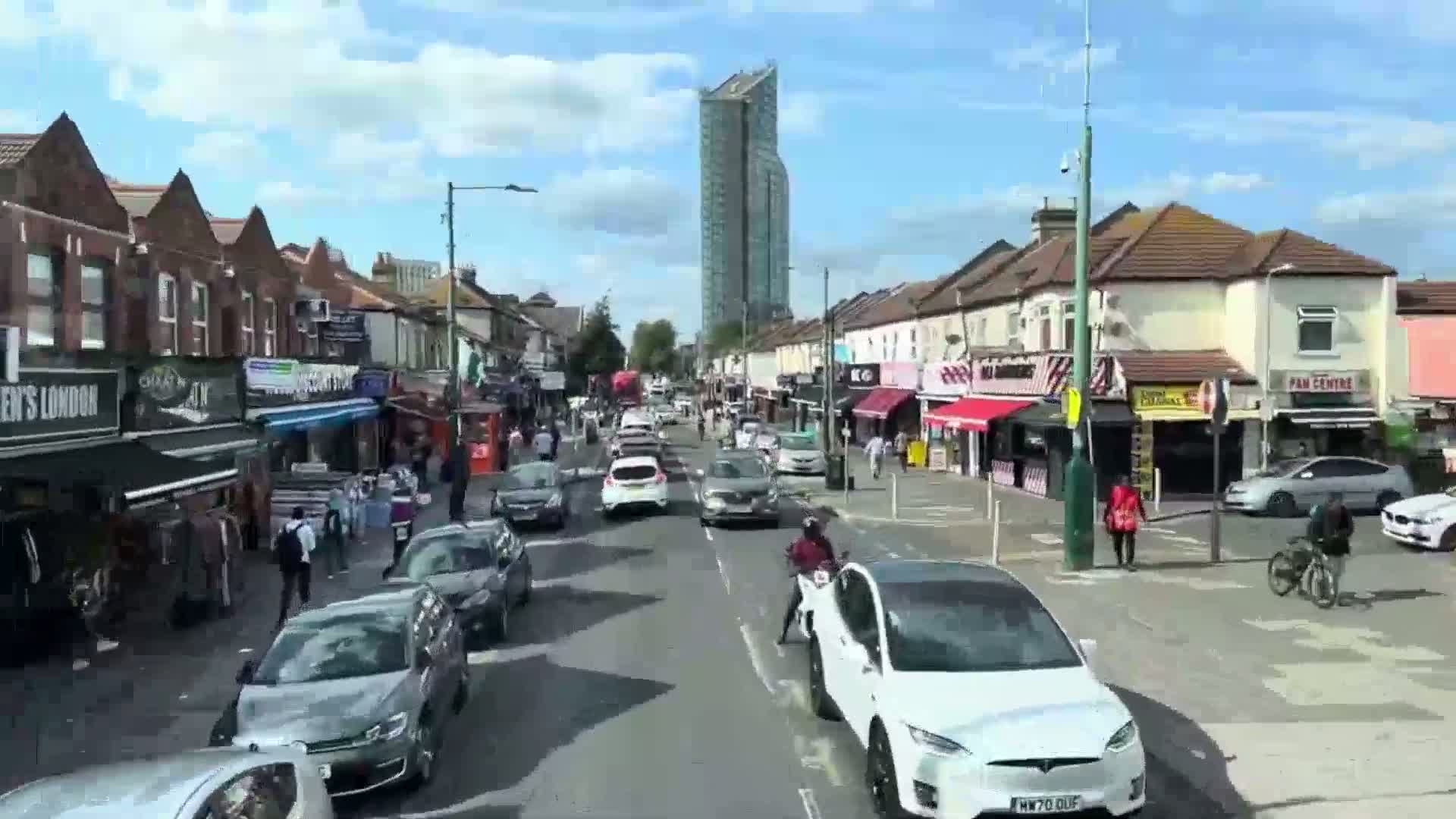 A sunny day reveals a bustling street scene with pedestrians and cars moving along a road lined with shops and residential buildings, a tall building visible in the background.