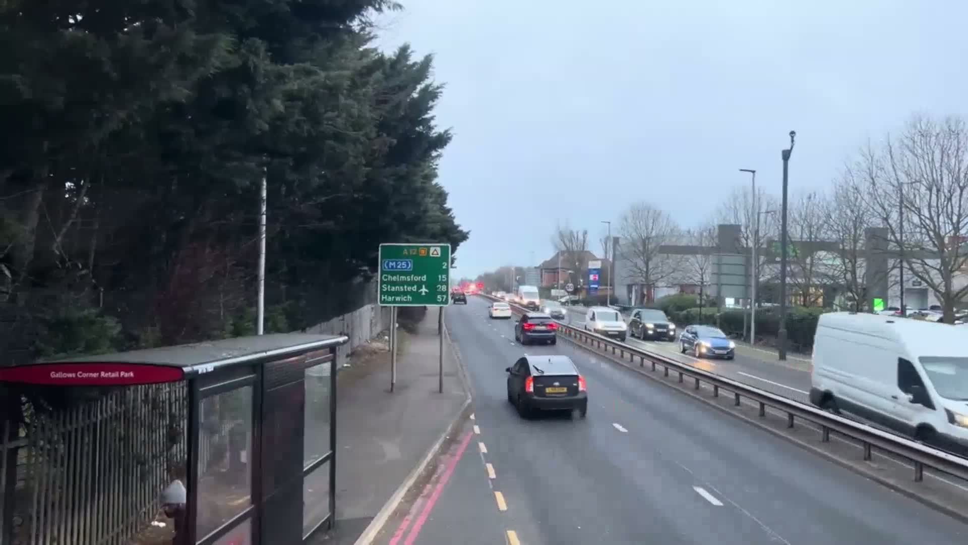 A multi-lane road with moderate traffic flows under an overcast sky, lined with trees and shrubs, alongside a bus stop.