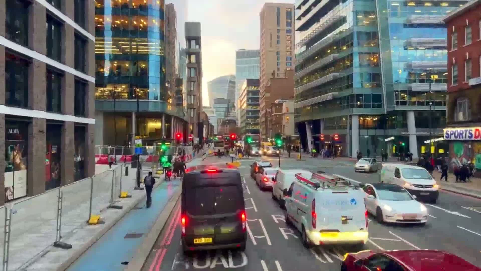 A city street scene at dusk shows modern buildings, pedestrians, and a variety of vehicles including vans, cars, and buses moving through an intersection.