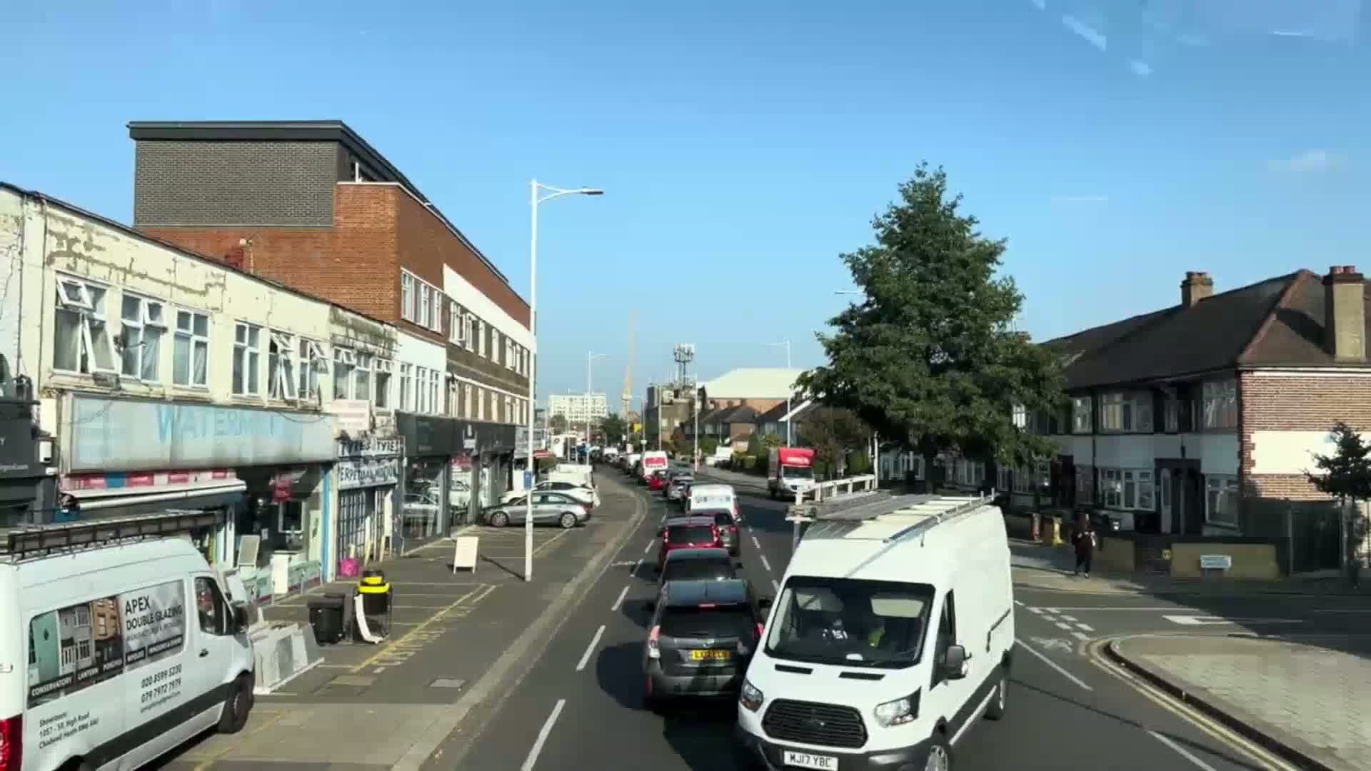 A sunny day reveals a street scene of parked cars and slow-moving traffic along a road lined with shops and residential buildings.
